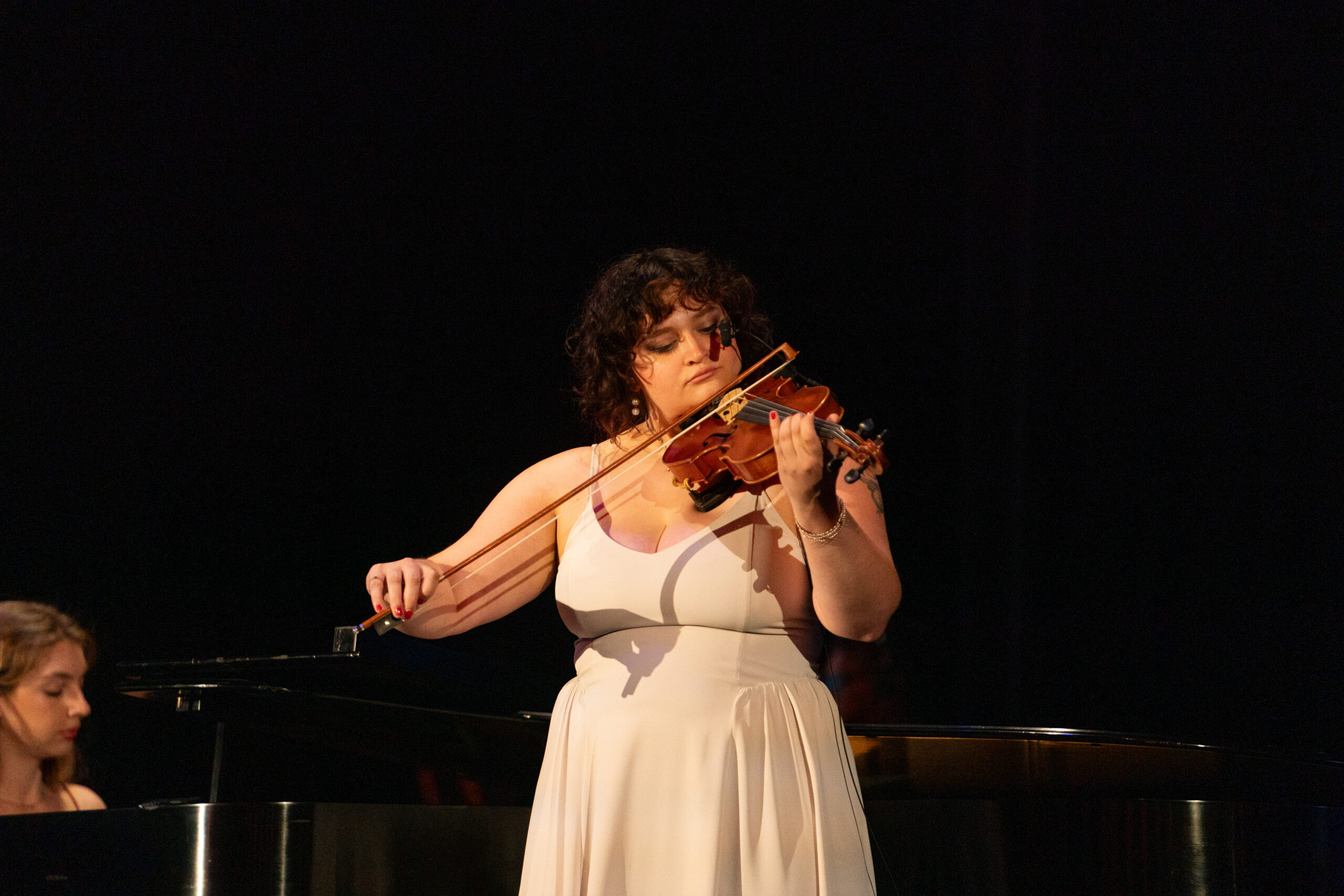 A violinist in a white dress performs on stage under dramatic lighting, with a pianist in the background.