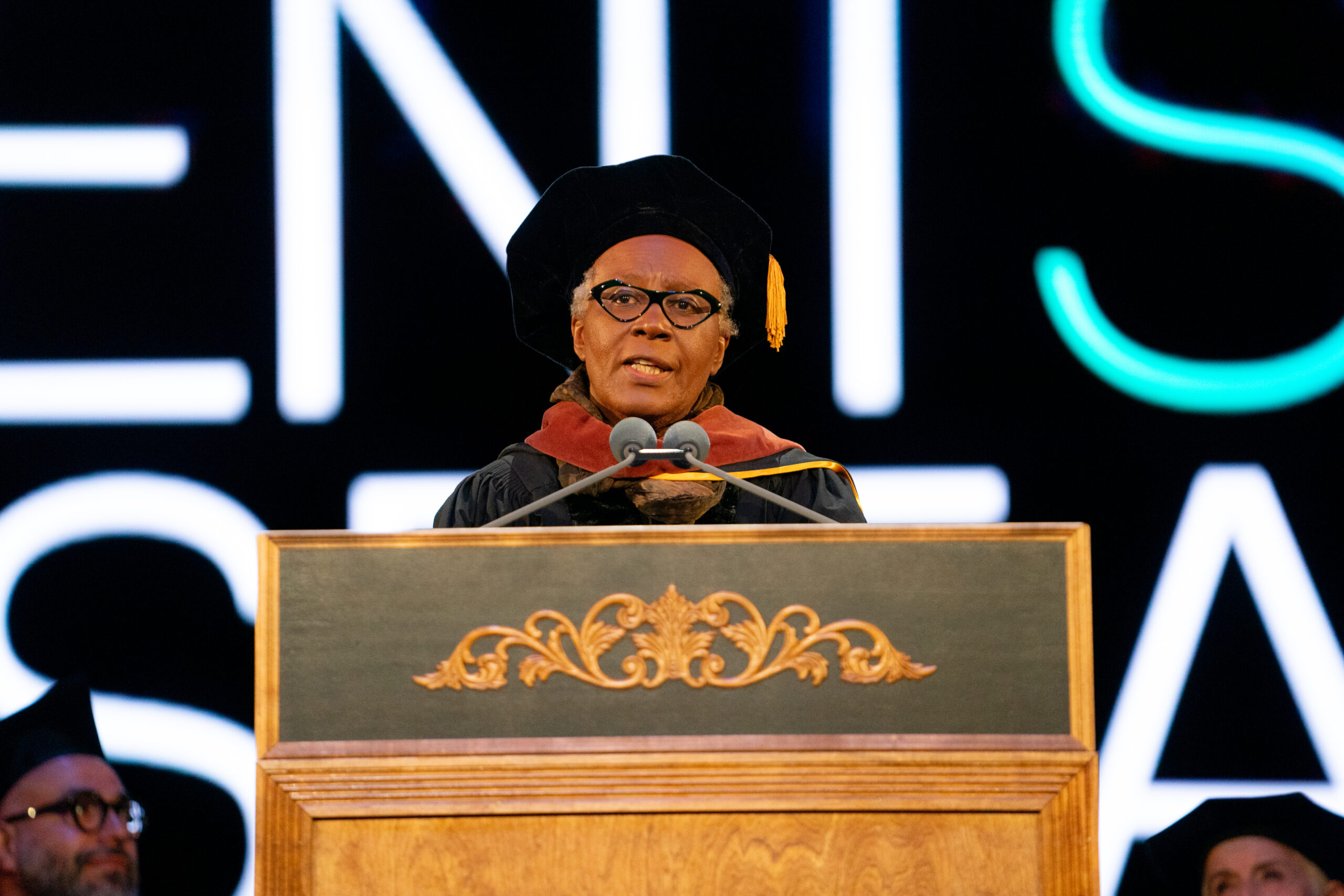 A commencement speaker in academic regalia addresses the audience from a podium, standing in front of a lit digital backdrop.