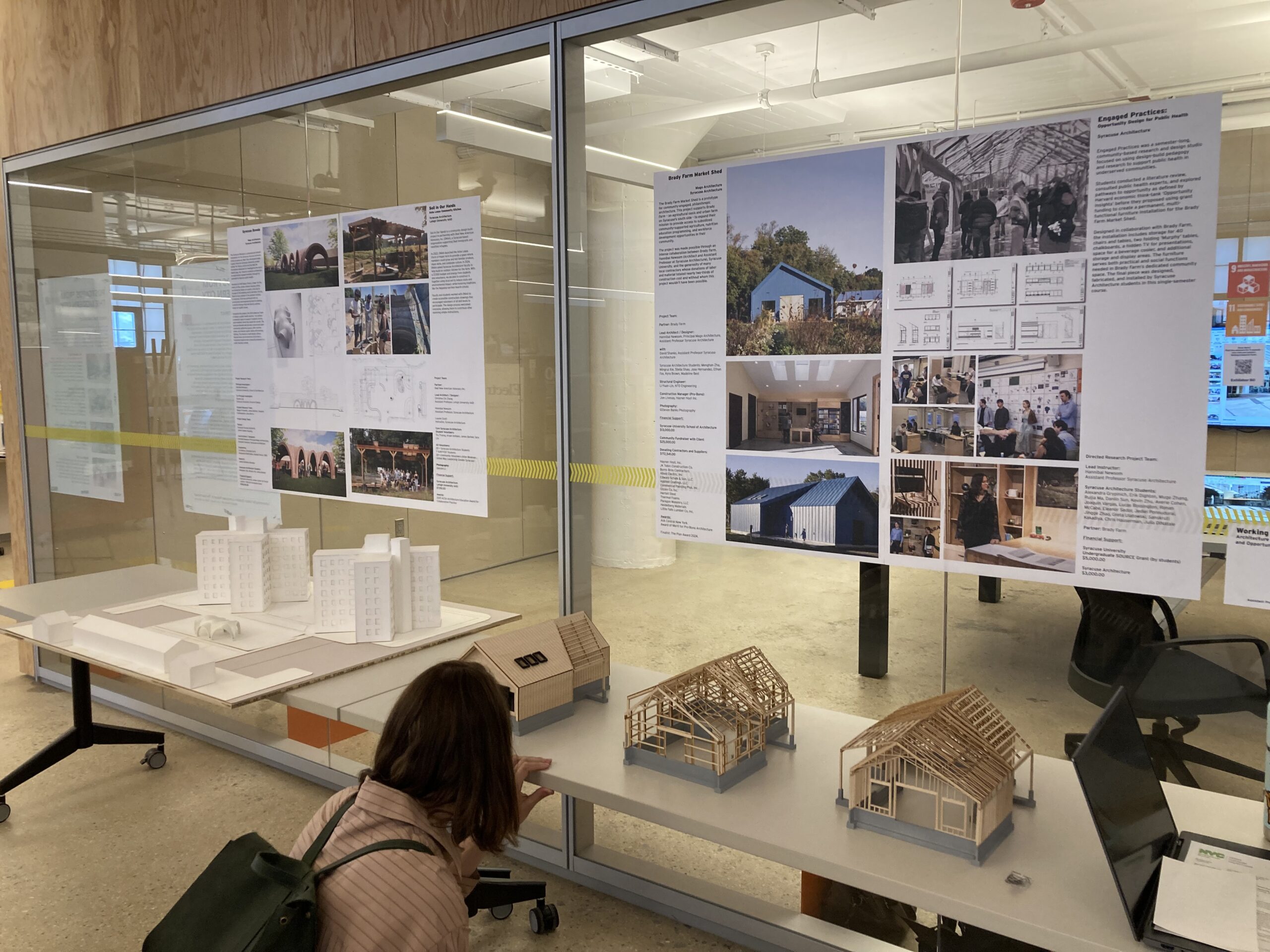 Interior of a university design studio displaying architectural posters and scale models. A student crouches in front of small wooden house models, while a large glass wall in the background features printed research boards detailing community-based housing projects.