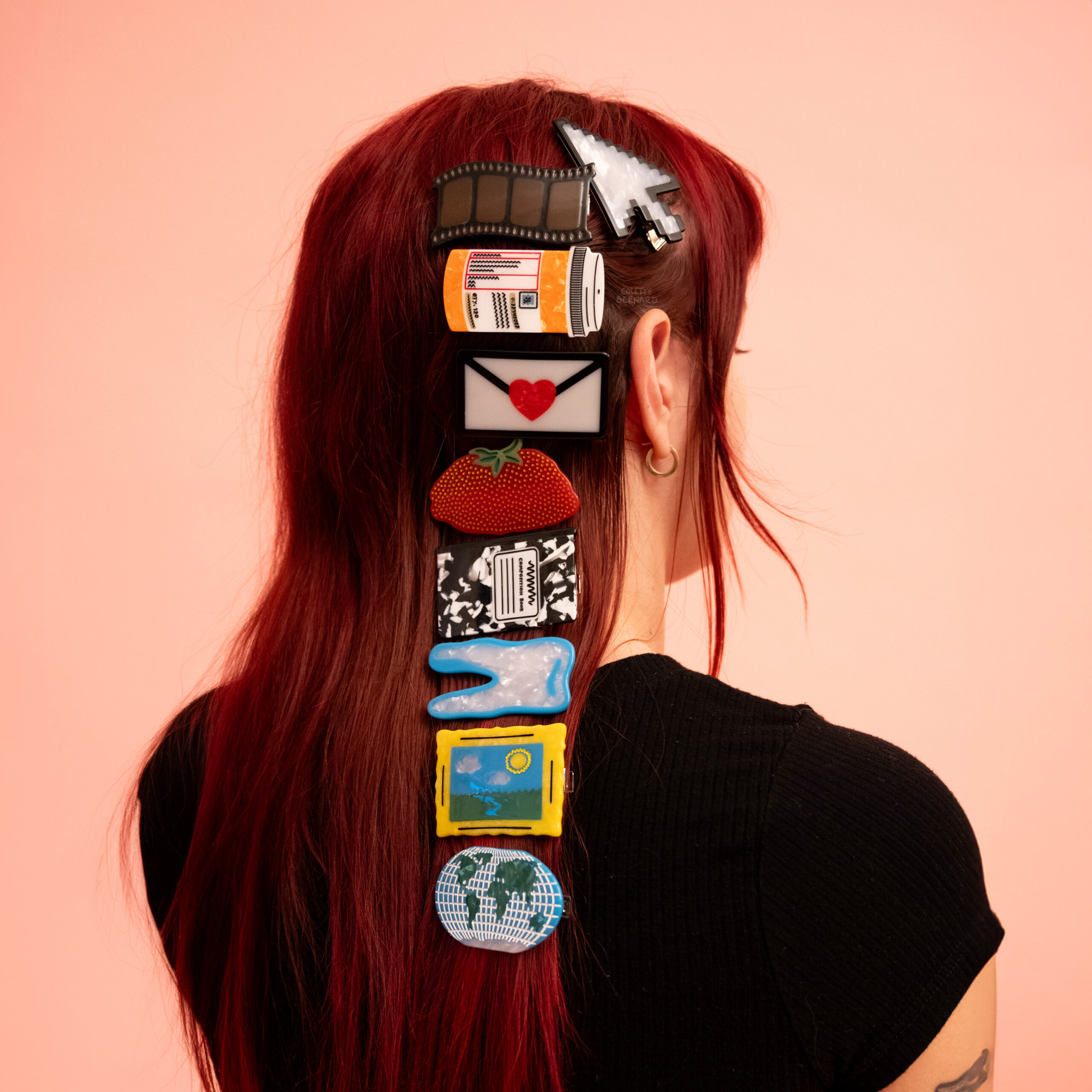 Hairclips worn in a row by by a model with red hair. From top to bottom, the clips are a cursor icon, film strip, pill bottle, envelope sealed with a heart, smashed strawberry, composition notebook, tooth, a painting, and a world map.