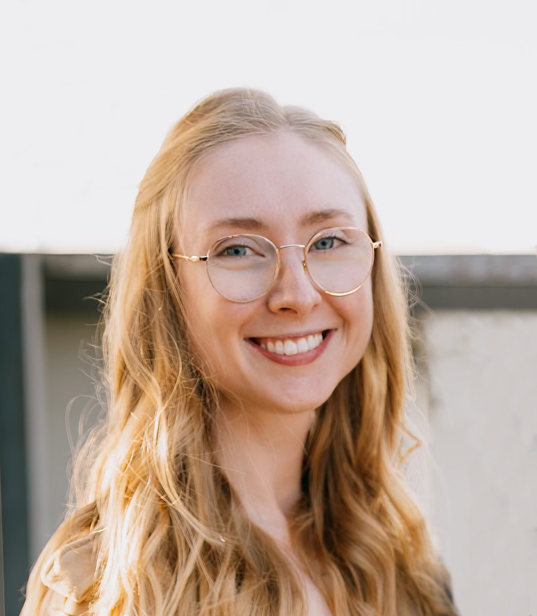 Headshot of Sydney Barton in natural light, wearing glasses and smiling at the camera