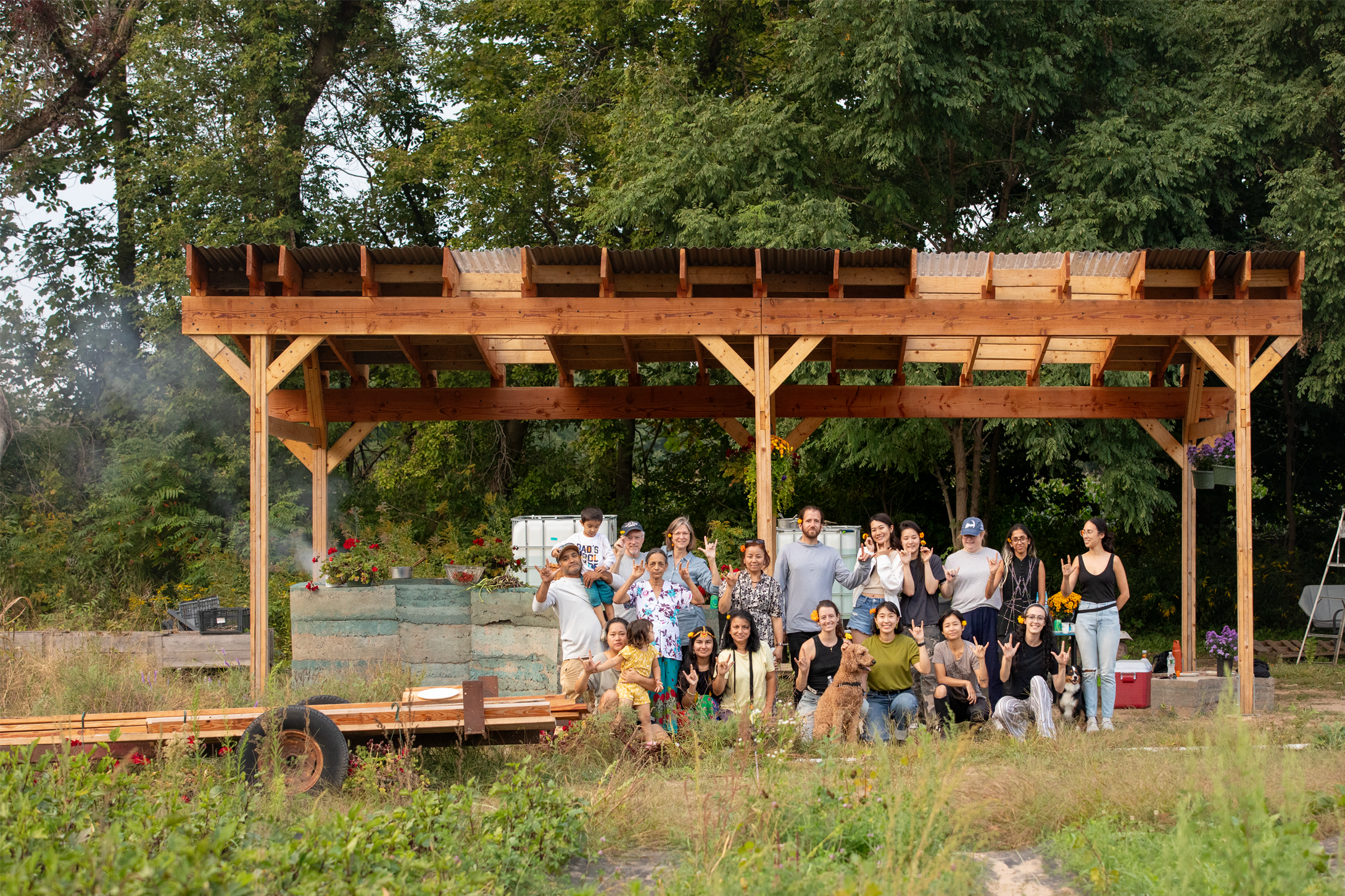A group of people posing in front of a newly built outdoor community kitchen structure made of timber with a corrugated metal roof. The group includes adults, children, and pets, standing on a grassy plot surrounded by trees and gardening elements, celebrating the project opening.