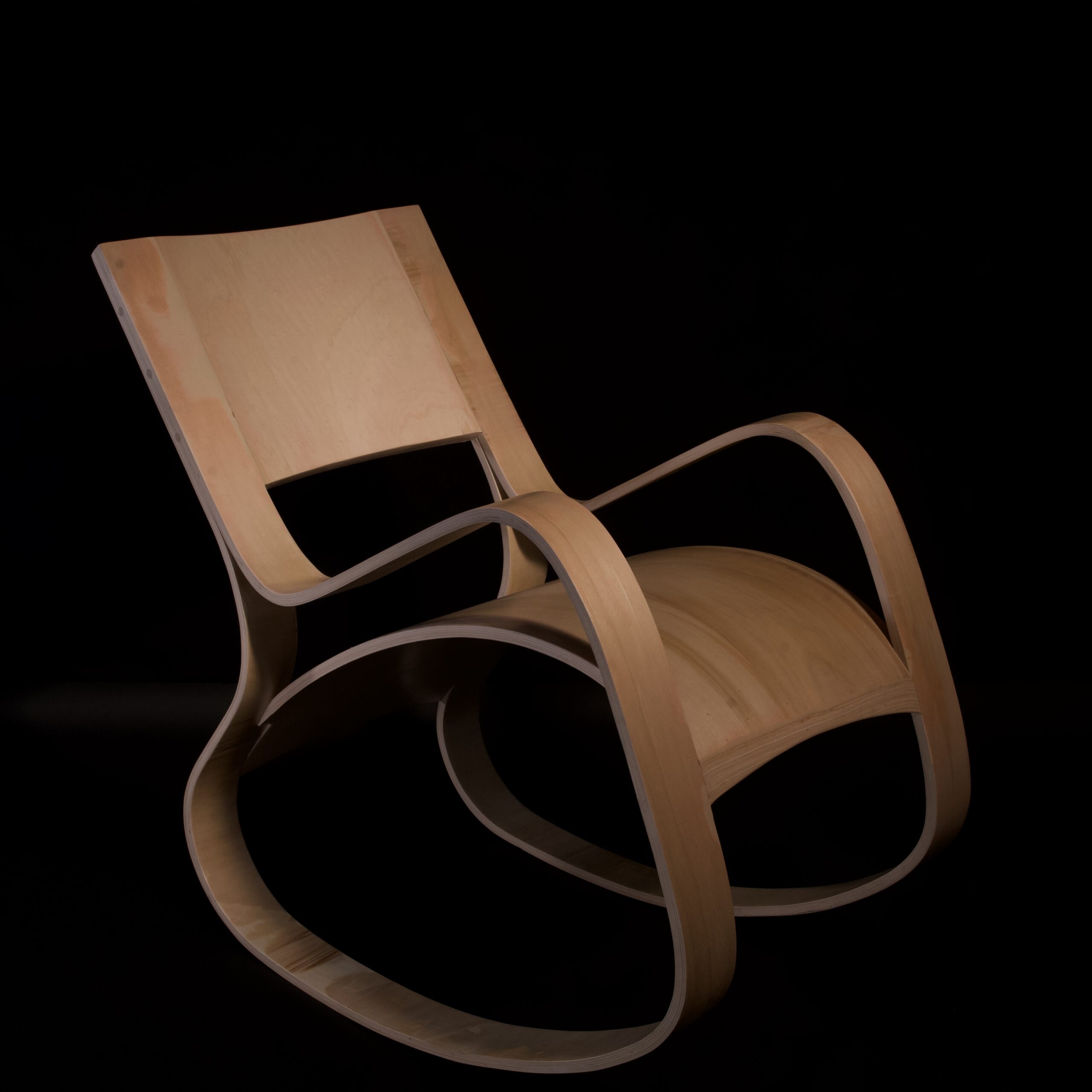A finished bent plywood rocking chair with a continuous loop frame and smooth, flowing curves. The chair is positioned in front of a black background, highlighting its sculptural form and craftsmanship in minimalist, modern design.