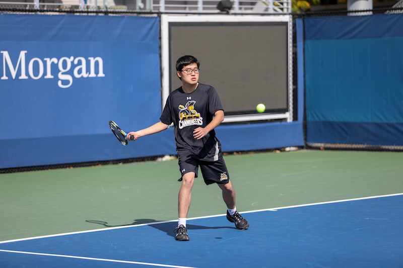 Student playing tennis on an outdoor court wearing “CANNONEERS” team gear.