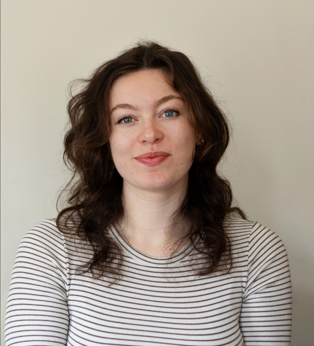 A headshot of a woman with long brown hair, looking at the camera. She stands in front of a white wall.