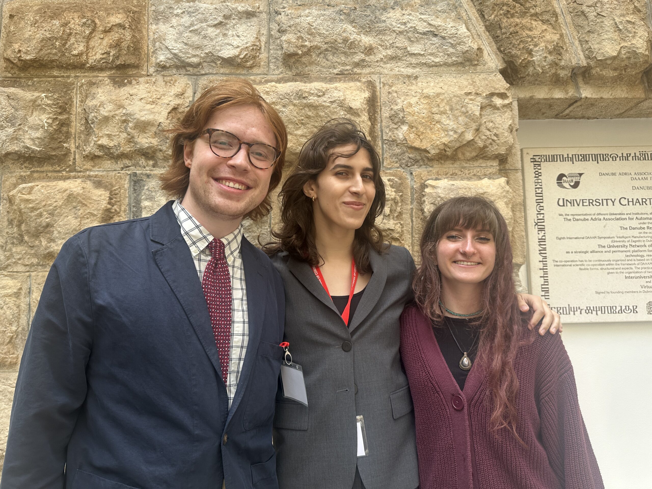 Three people smiling and posing together in front of a stone wall and university charter sign.