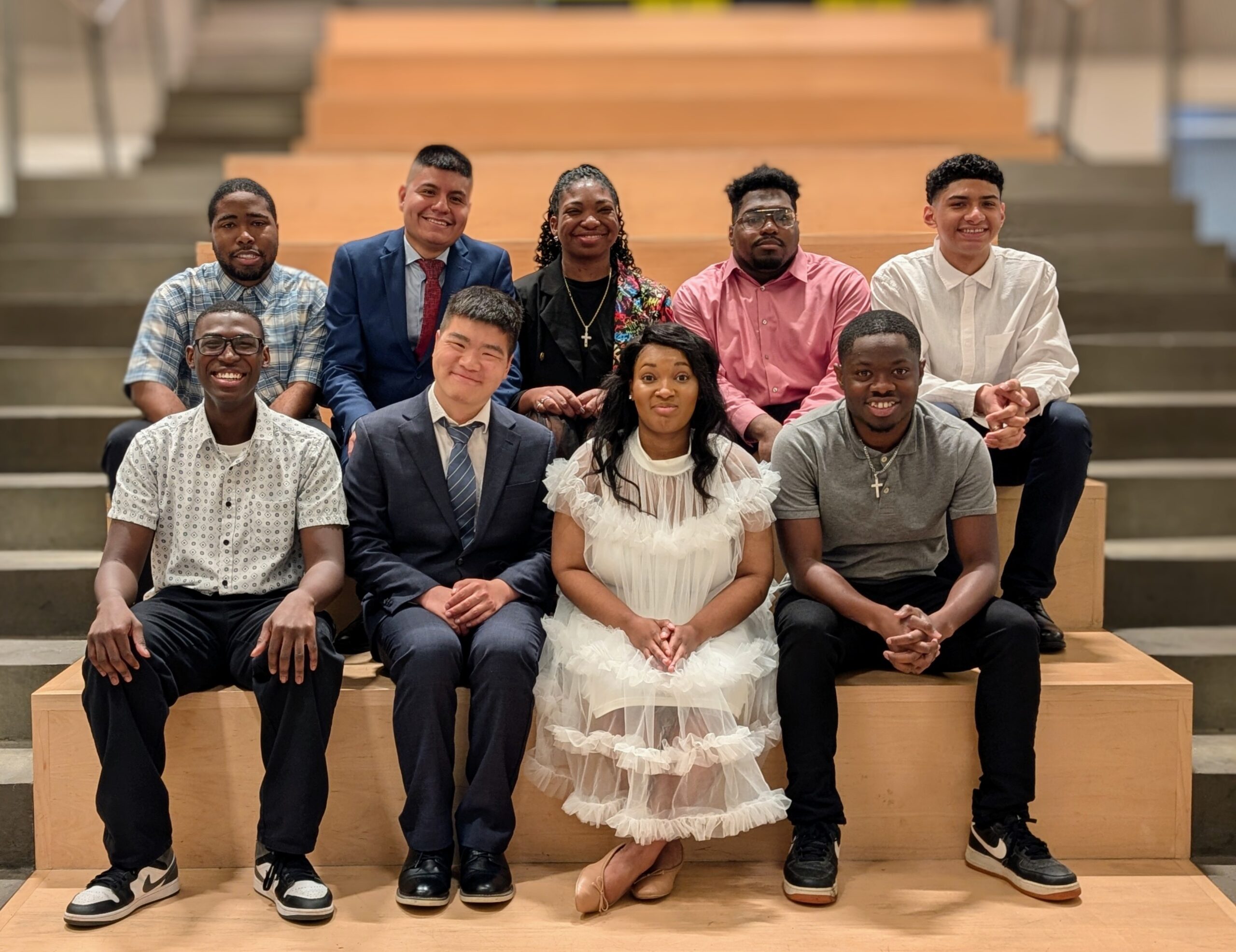 A group portrait of nine smiling Project SEARCH interns dressed in formal and semi-formal attire, seated together on wooden steps in a brightly lit interior space. The group includes a diverse mix of individuals, with some in suits, button-down shirts, and one wearing a white ruffled dress. They appear proud and celebratory, possibly marking their graduation or completion of the program.