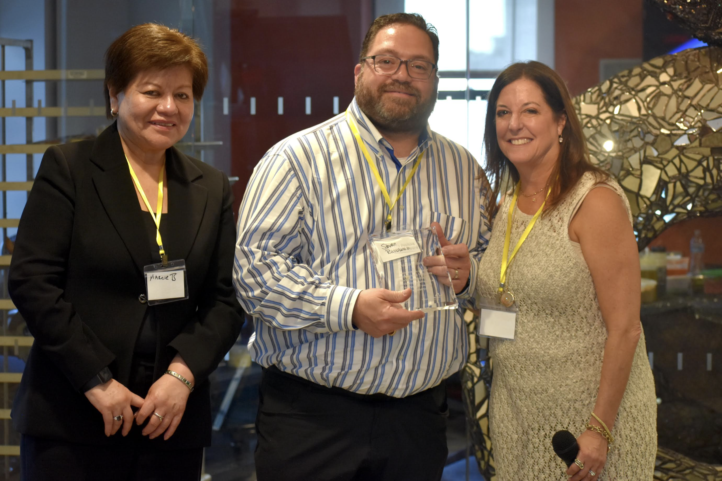 Three individuals pose together at an indoor event. The person in the center, wearing a striped shirt and glasses, smiles while holding a clear glass award. To the left stands a person in a black suit with a name tag that reads “Maric B.” On the right, a woman in a sleeveless cream dress holds a microphone and smiles. All three wear yellow lanyards. A mosaic sculpture is visible in the background.