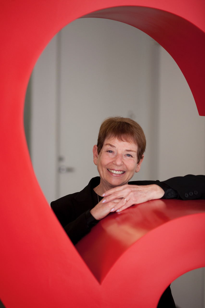 Alisa Zamir smiles leaning against and peering through a large red curved sculpture