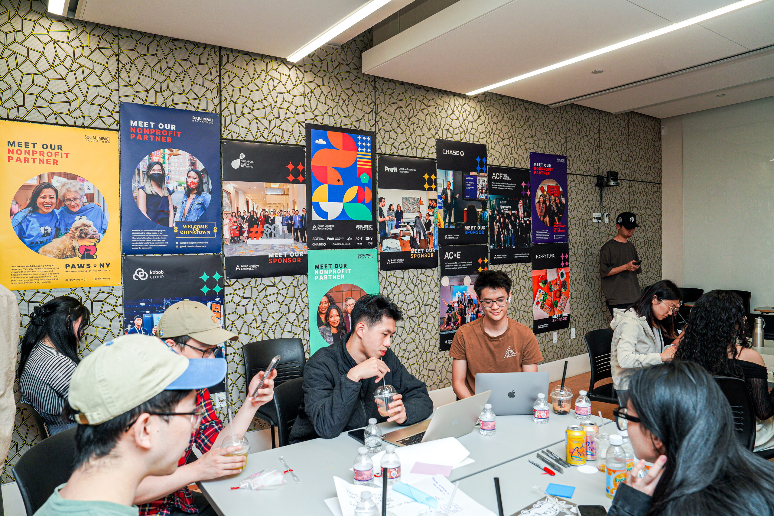 People sitting around tables with laptops and pens and waters, working together on a project. In the background are colorful posters featuring partners and sponsors for the event.