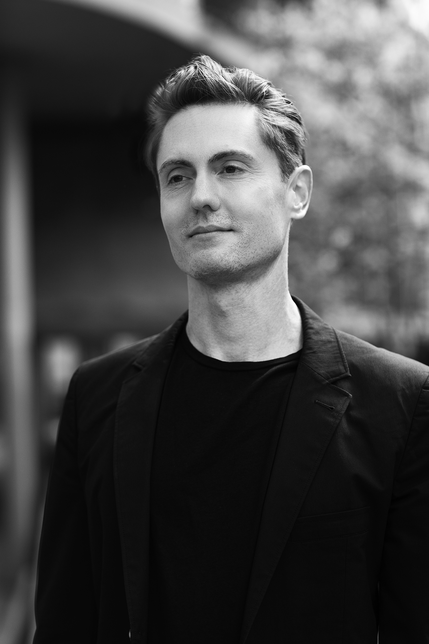 A black and white portrait of a man standing outdoors, dressed in a black blazer over a black T-shirt. He has short, neatly styled hair and a calm, confident expression, gazing slightly to the side. The background is softly blurred, showing hints of modern architecture and foliage.