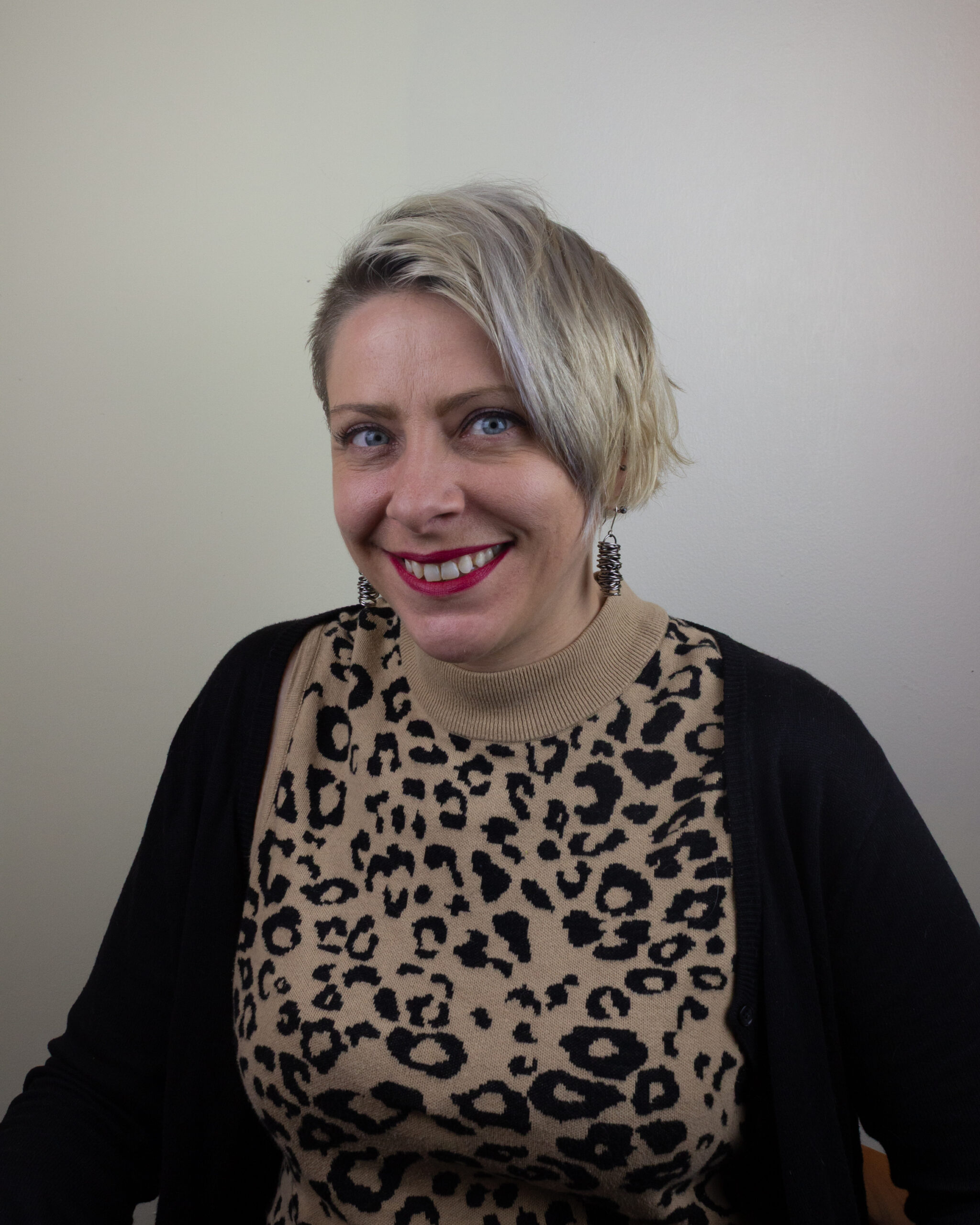 A woman with short platinum blonde hair and a side part smiles while seated in front of a neutral wall. She is wearing a tan sweater with a black leopard print, a black cardigan, and statement earrings.