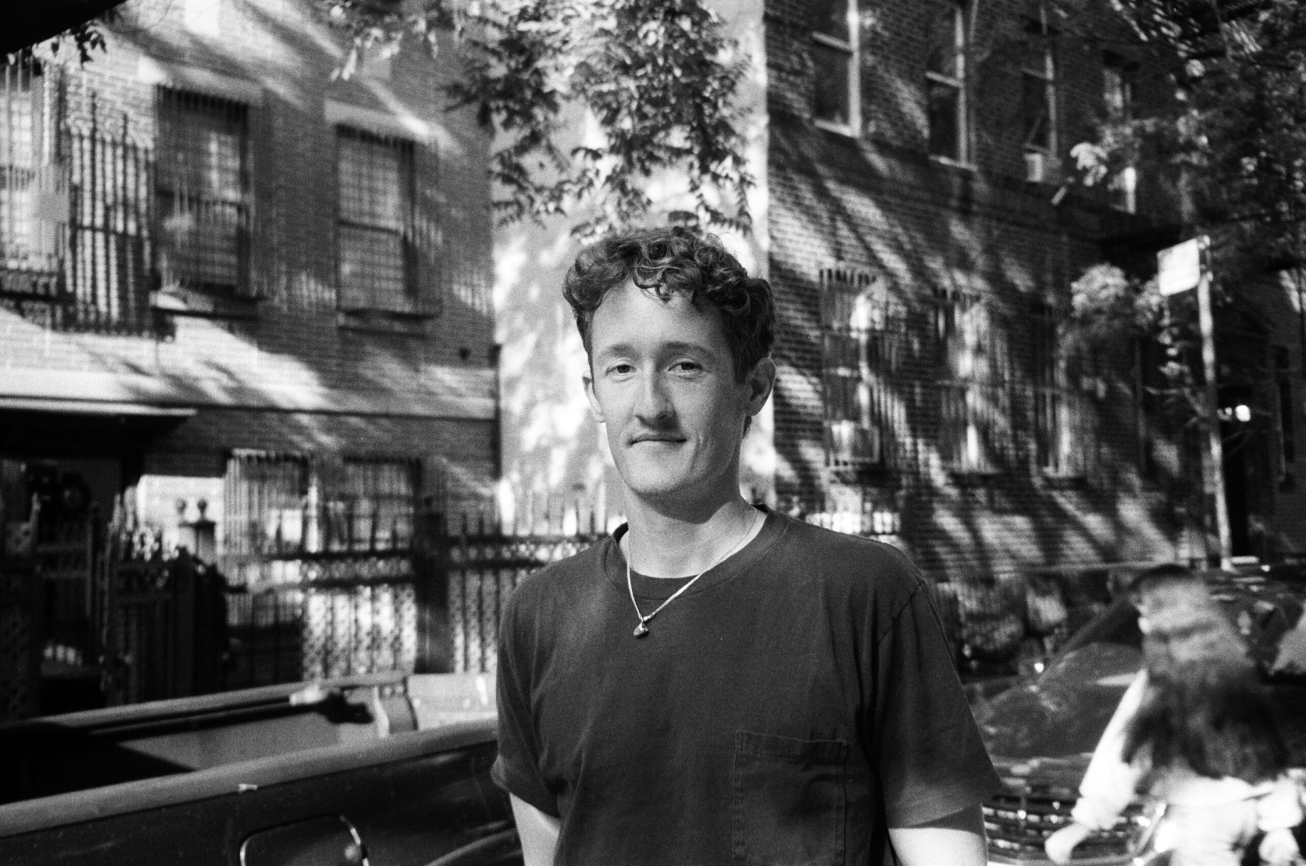 A black and white street portrait of a man standing outdoors in front of a residential brick building with barred windows and wrought iron fencing. He wears a dark T-shirt and a simple necklace, looking directly at the camera with a slight smile. Dappled sunlight and tree shadows fall across the wall behind him. In the foreground, part of a parked car is visible, and a blurred pedestrian passes by on the right edge of the frame.