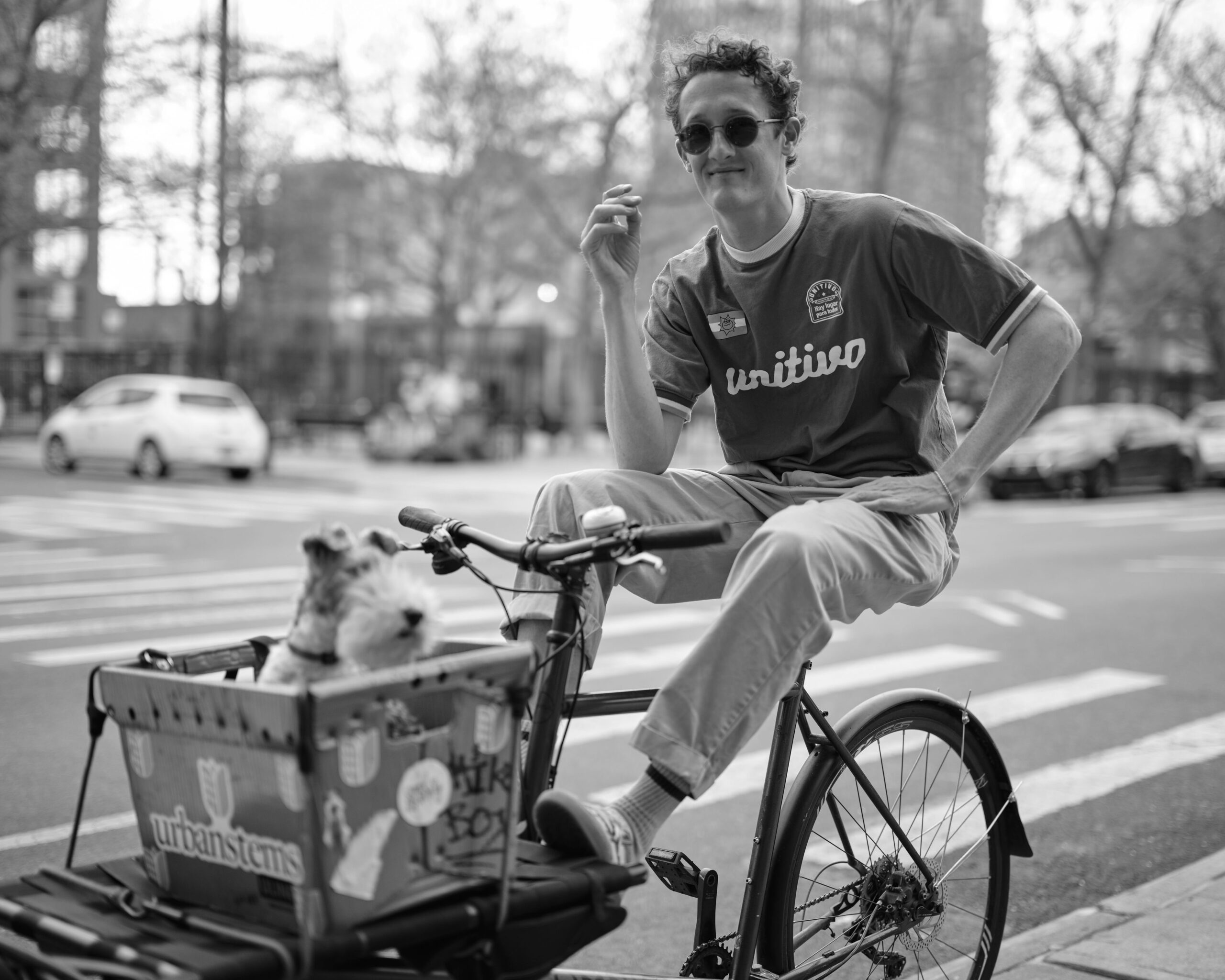 A black and white photo of a man sitting casually on a bicycle stopped at a city intersection. He wears sunglasses, a short-sleeved “unitivo” jersey-style shirt, and cuffed light pants. One hand rests on the handlebars while the other is lifted mid-gesture. A small dog with pointed ears sits in a front-mounted crate labeled “urbanstems,” which is decorated with various stickers. Parked cars and a blurred cityscape with trees and tall buildings fill the background.