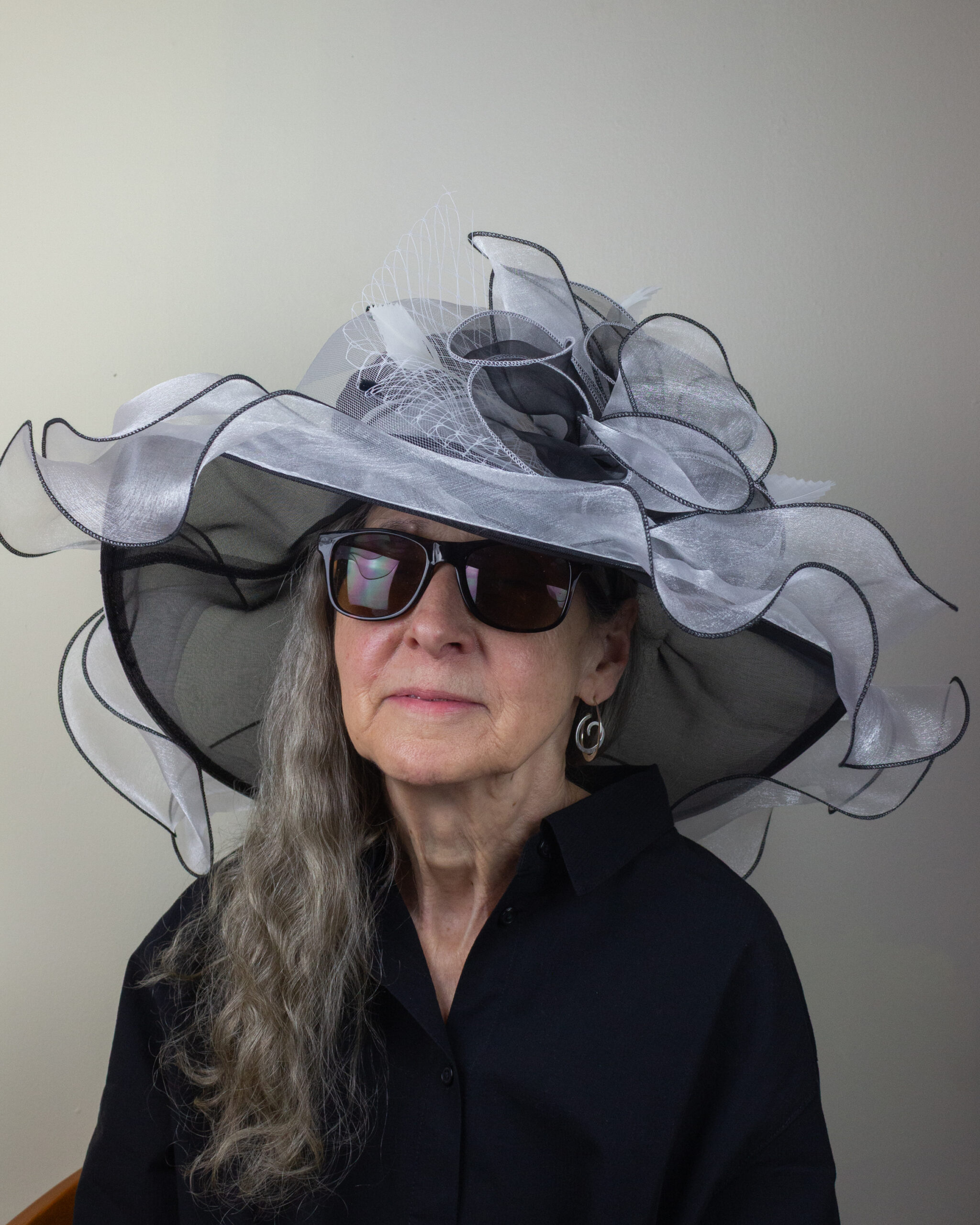 An older woman with long gray hair wears oversized black sunglasses and a large, decorative black and white hat with mesh and ribbon details. She is dressed in a black button-up shirt and is seated against a plain background.