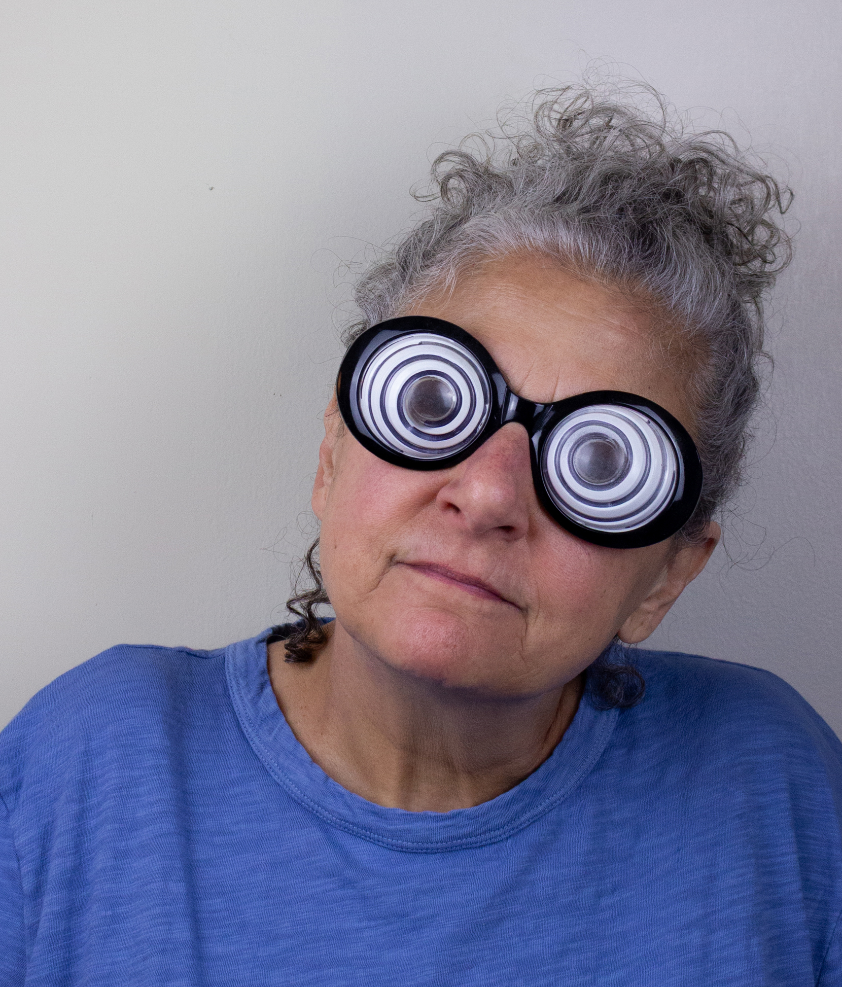 A woman with curly gray hair pulled back wears a bright blue shirt and large novelty spiral glasses that create a hypnotic effect. She has a neutral expression and is posed against a light-colored wall.