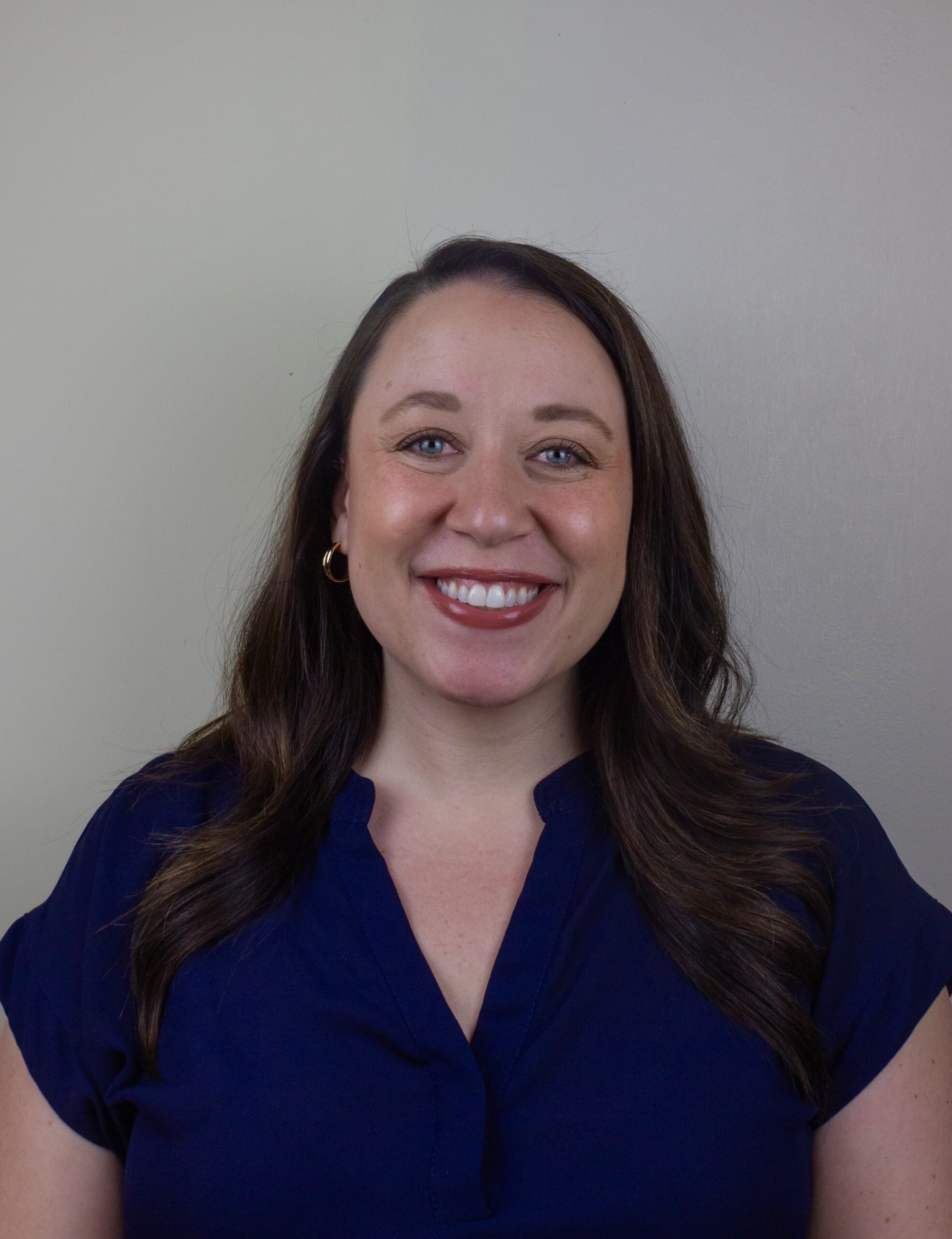 A woman with long brown hair styled in loose waves smiles warmly. She is wearing a navy blue blouse with gold hoop earrings and is posed in front of a plain light gray wall.
