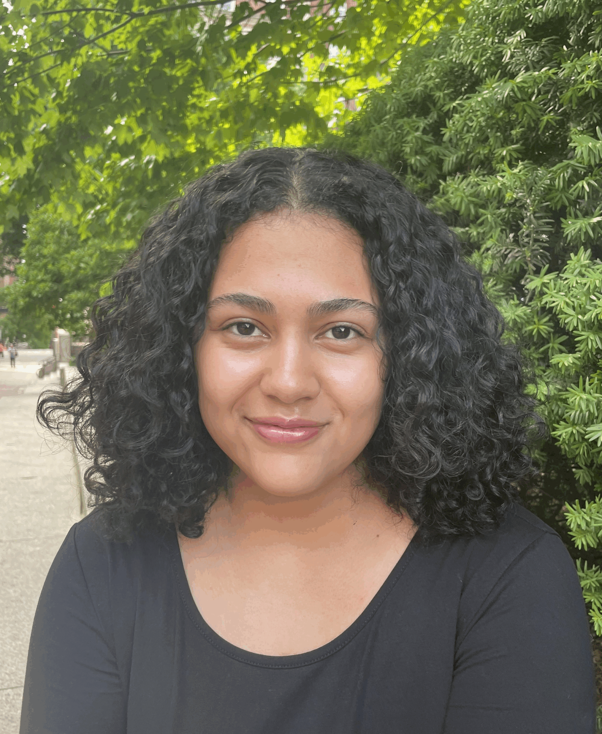 A woman with curly black hair smiles gently while sitting outdoors. She is wearing a black top, and leafy green trees and a building are visible in the background.