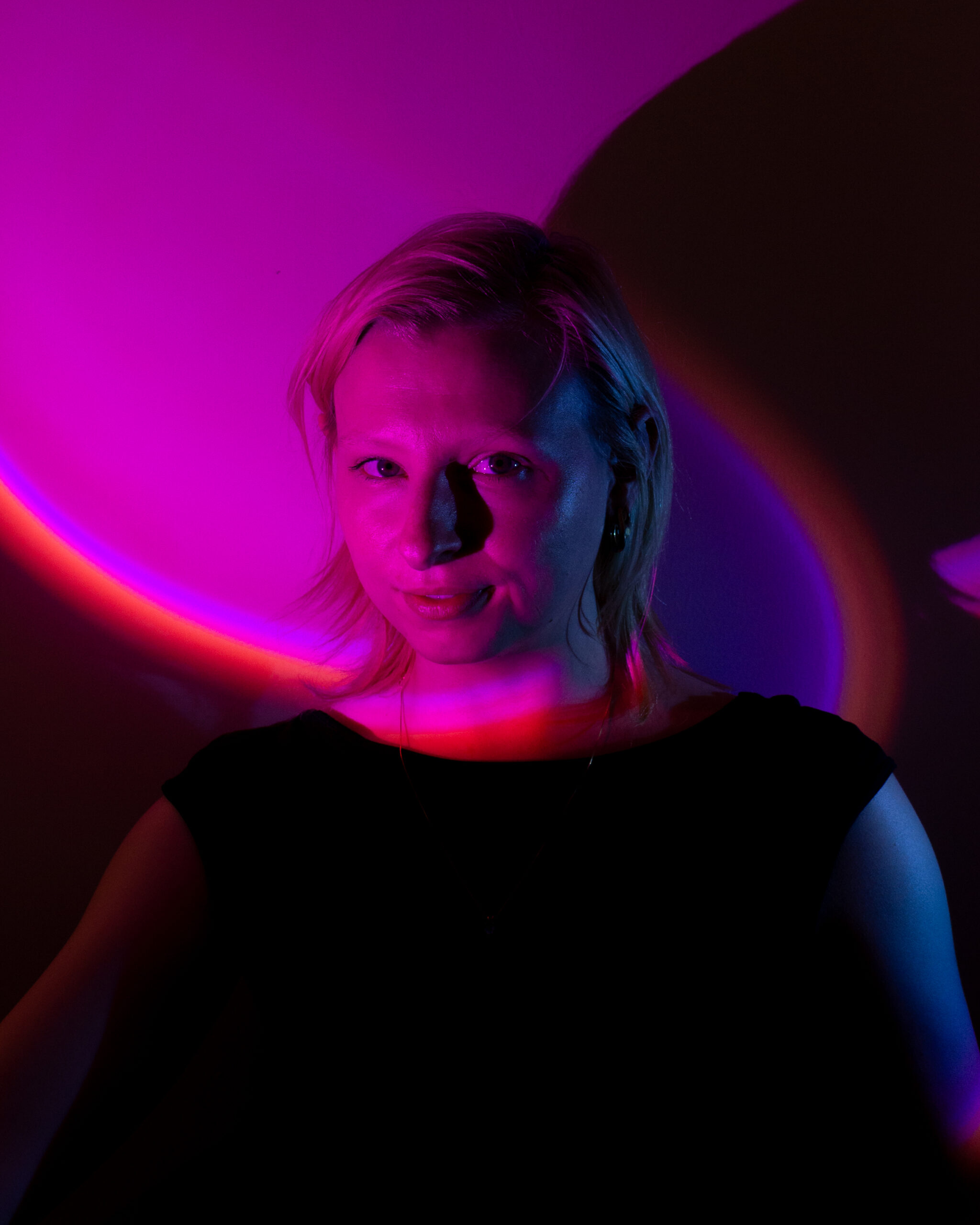 A woman with short blonde hair is lit dramatically with pink and purple lights against a dark background. She is wearing a black top and a subtle necklace, with vibrant lighting curving across her face and shoulders.