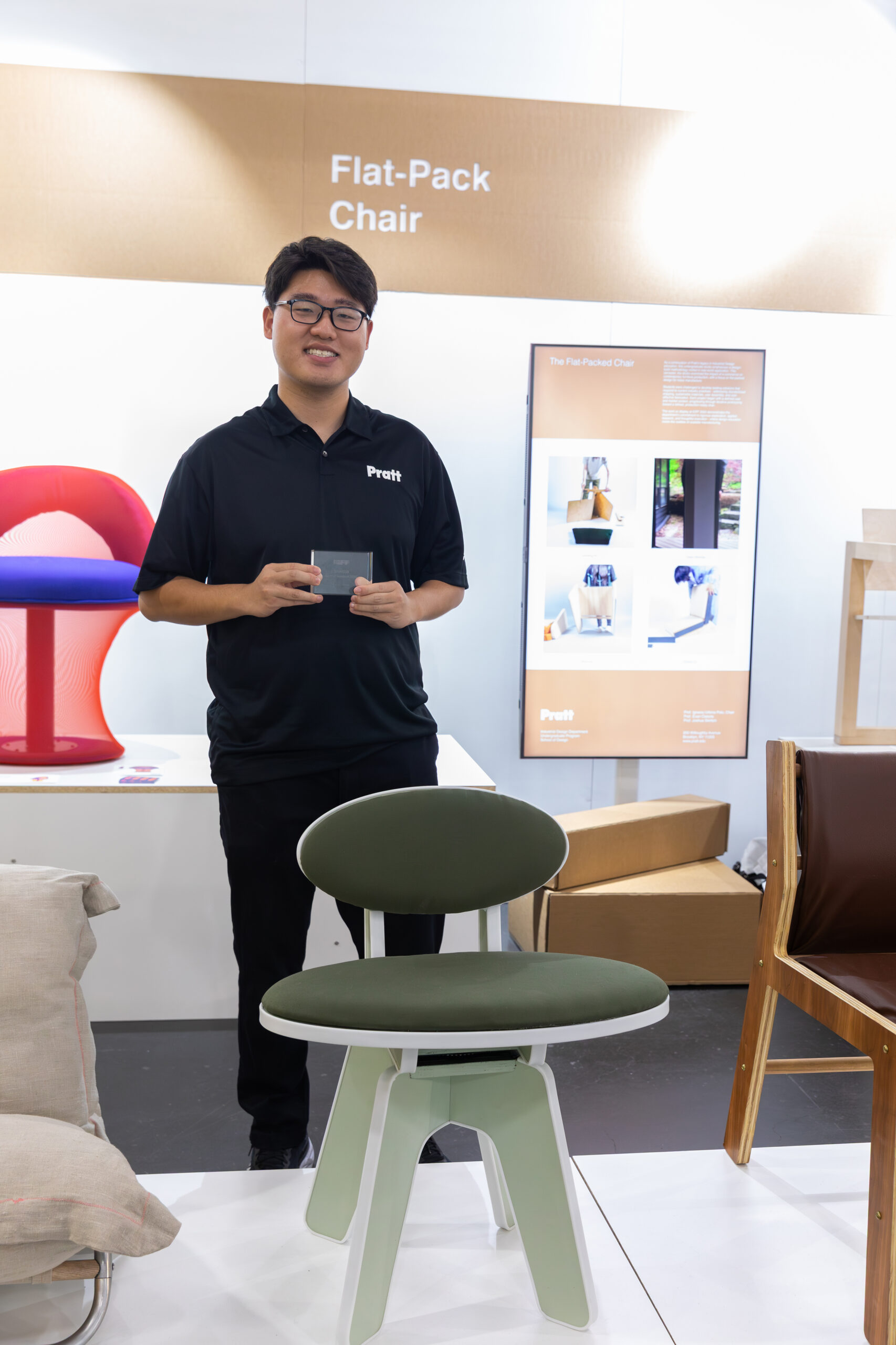 A young man wearing a black "Pratt" polo shirt stands smiling in front of a design exhibit labeled "Flat-Pack Chair." He holds a small clear award or plaque. In front of him is a modern green and white chair with a circular seat and backrest. Behind him are display screens and other design pieces, including a red and purple chair and a brown wood-framed chair. The exhibit appears to be in a well-lit showroom or gallery space.