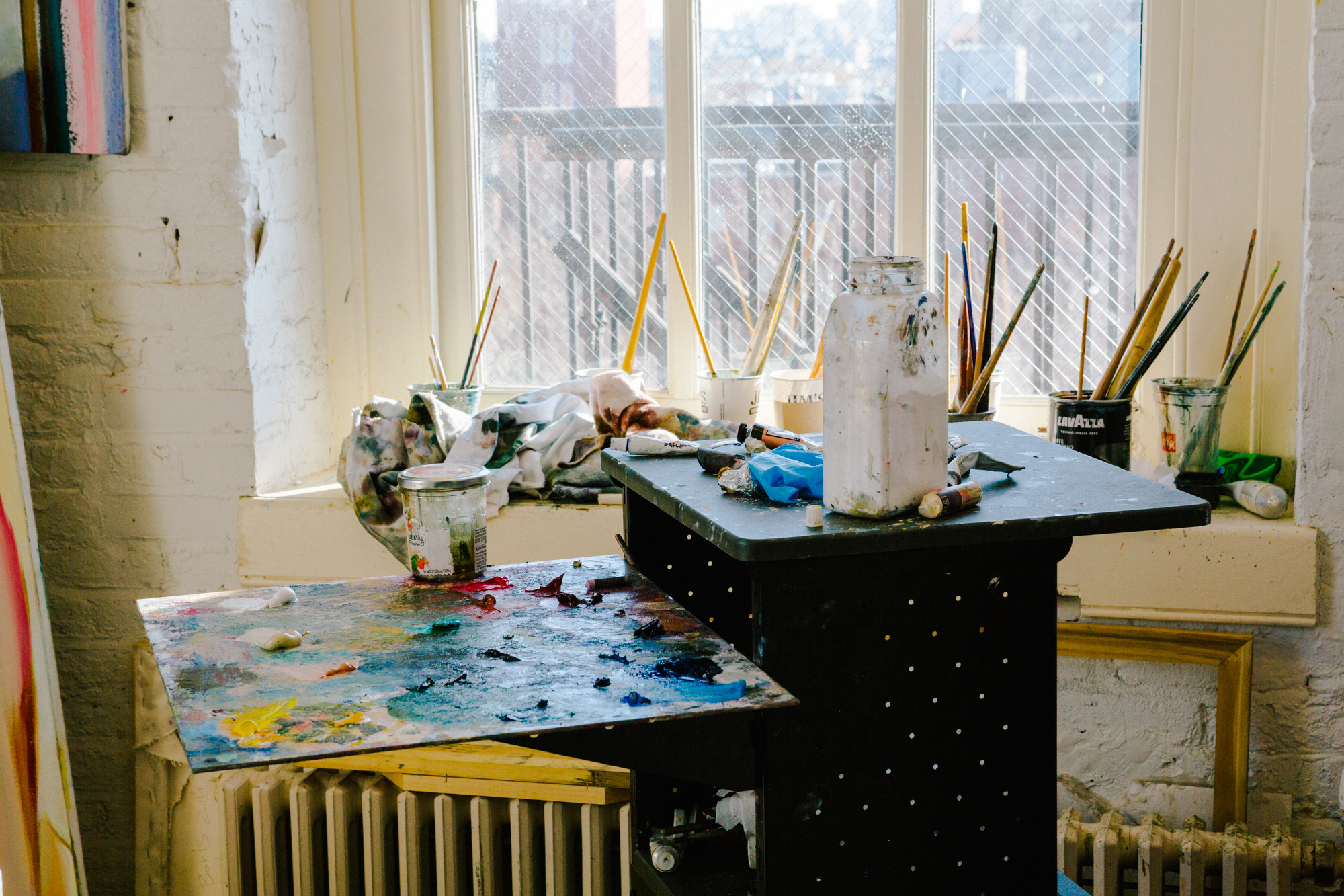 An artist’s studio corner with a sunlit window behind a cluttered work area. Multiple jars and containers holding paintbrushes are lined up on the windowsill. The table in the foreground is covered with tubes of paint, a stained palette with vibrant smears of color, and a large white paint container. A radiator and painter’s rags sit below the window, with a wire-mesh screen visible outside. The overall atmosphere is warm, lived-in, and creatively active.