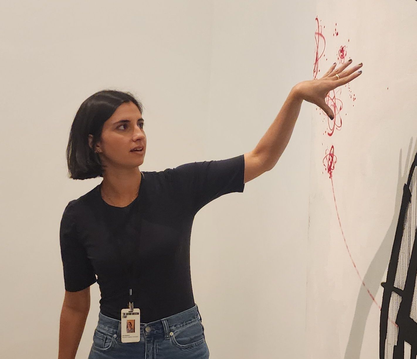 A woman in a black t-shirt and wearing an ID badge around her neck gestures at artwork a wall.