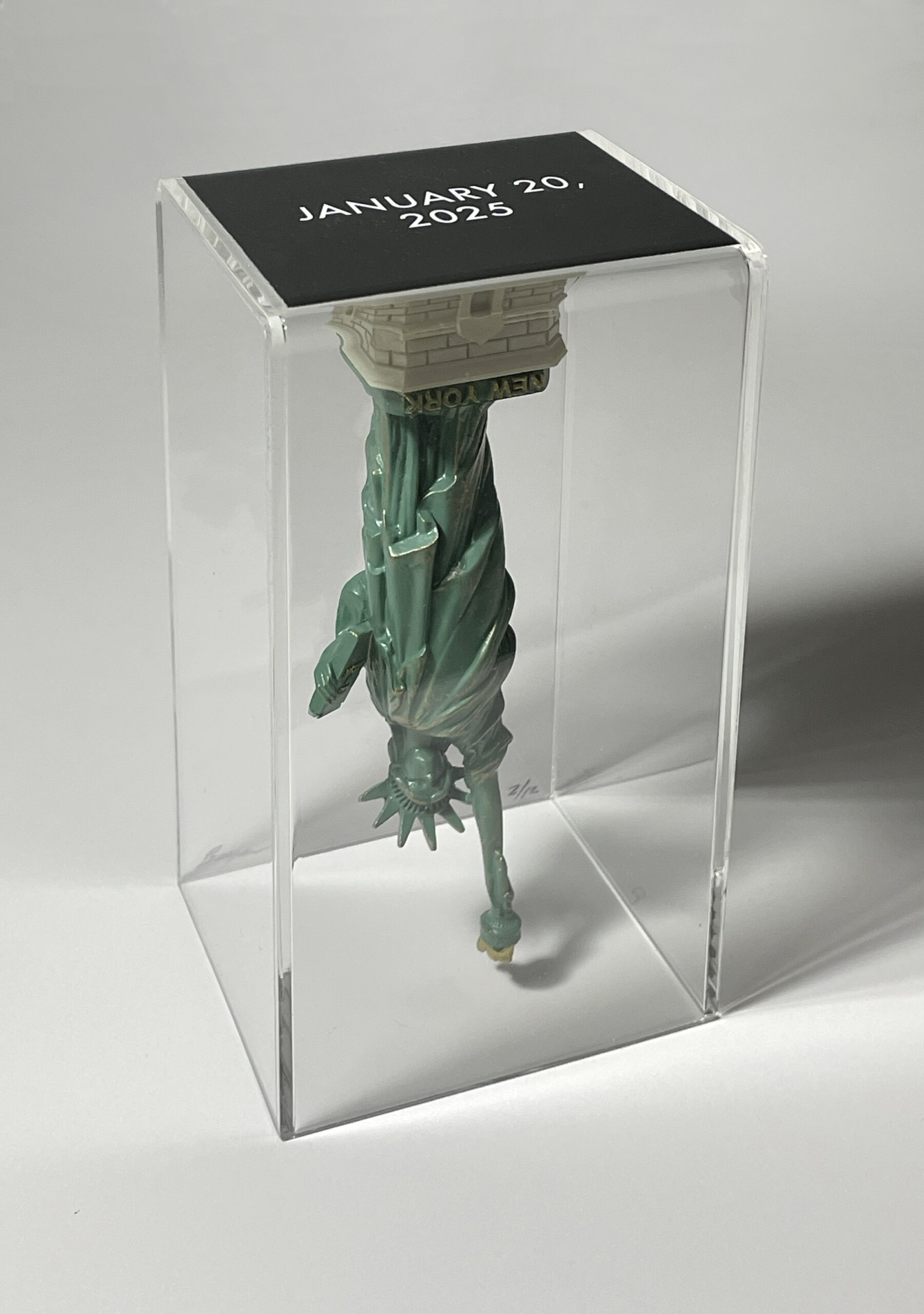 A small figurine of the Statue of Liberty is displayed upside down inside a clear acrylic box. The base of the statue, labeled "NEW YORK," is at the top, while the torch and crown point downward. The top panel of the box is black with white text that reads "JANUARY 20, 2025." The setup appears minimal and intentional, suggesting a symbolic or conceptual art piece.