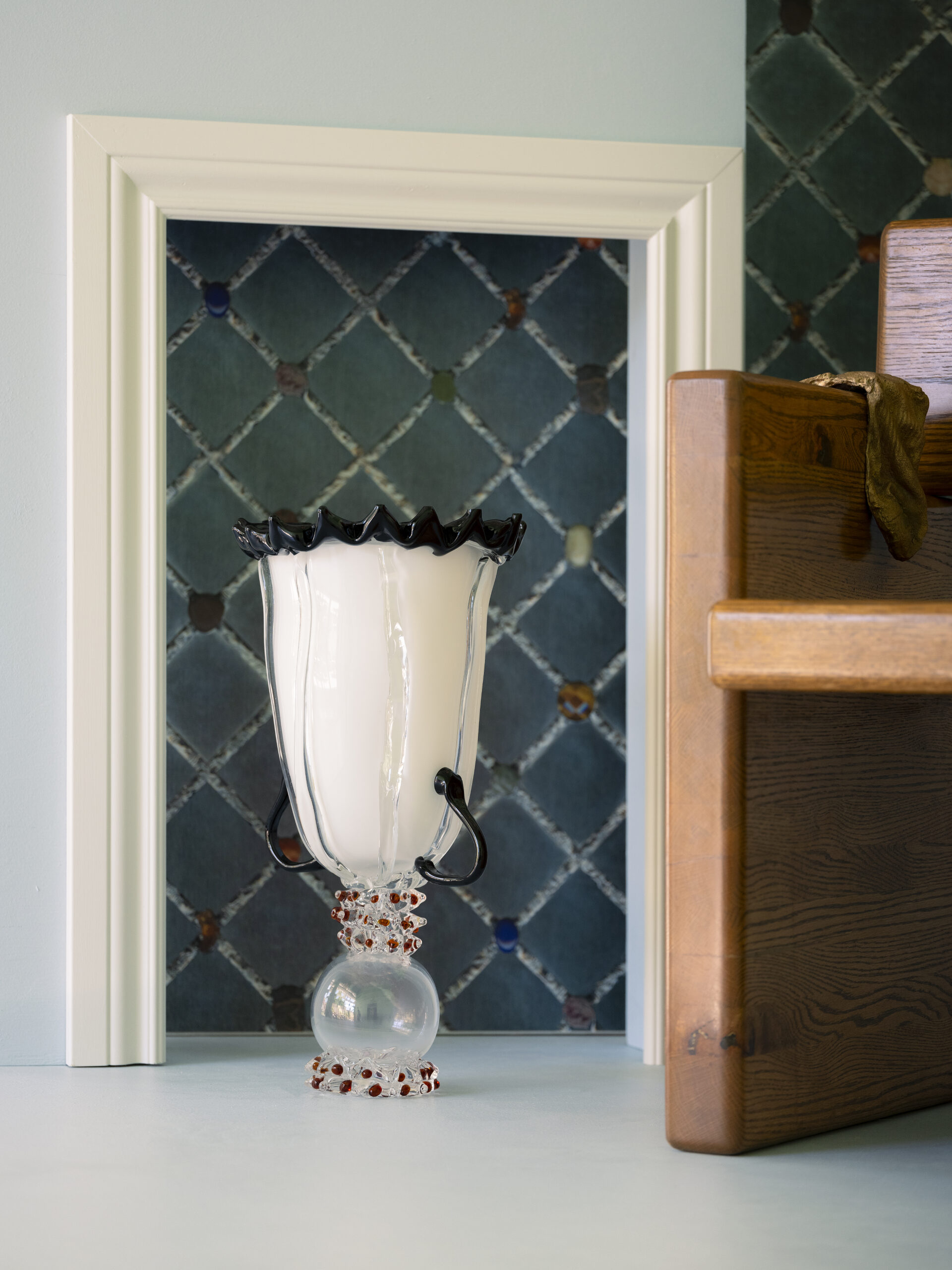 A decorative white glass vase with a wavy black rim and ornate clear base featuring red accents is displayed on a shelf. The vase is positioned inside a small, framed wall niche with a blue-green diamond-patterned tile backdrop. Part of a wooden piece of furniture is visible to the right, draped with a greenish-brown fabric. The scene is softly lit and artfully composed.