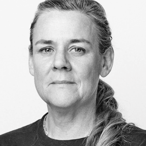 Black-and-white portrait of a woman with her hair pulled back in a low ponytail, wearing a simple dark top. She looks directly at the camera with a serious, steady expression, set against a plain white background.