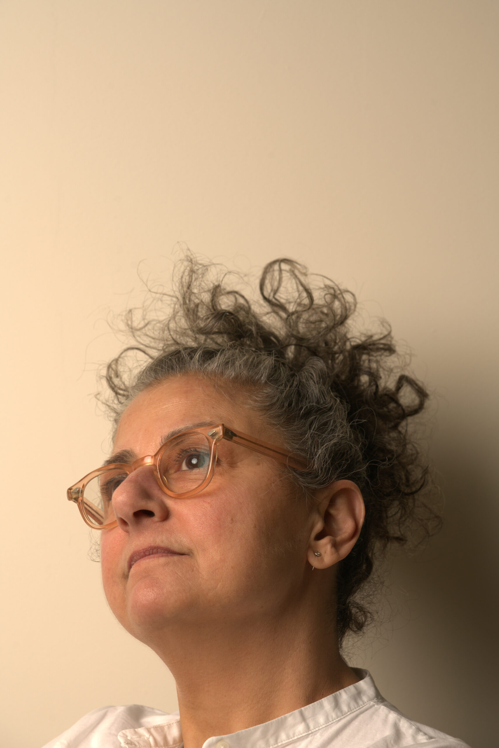 A close-up portrait of a person with salt-and-pepper curly hair styled upward, wearing light translucent glasses and a white button-down shirt. The subject gazes upward and to the left with a contemplative expression. The background is a smooth, beige wall, and the lighting is soft and natural, creating gentle shadows and a warm tone.