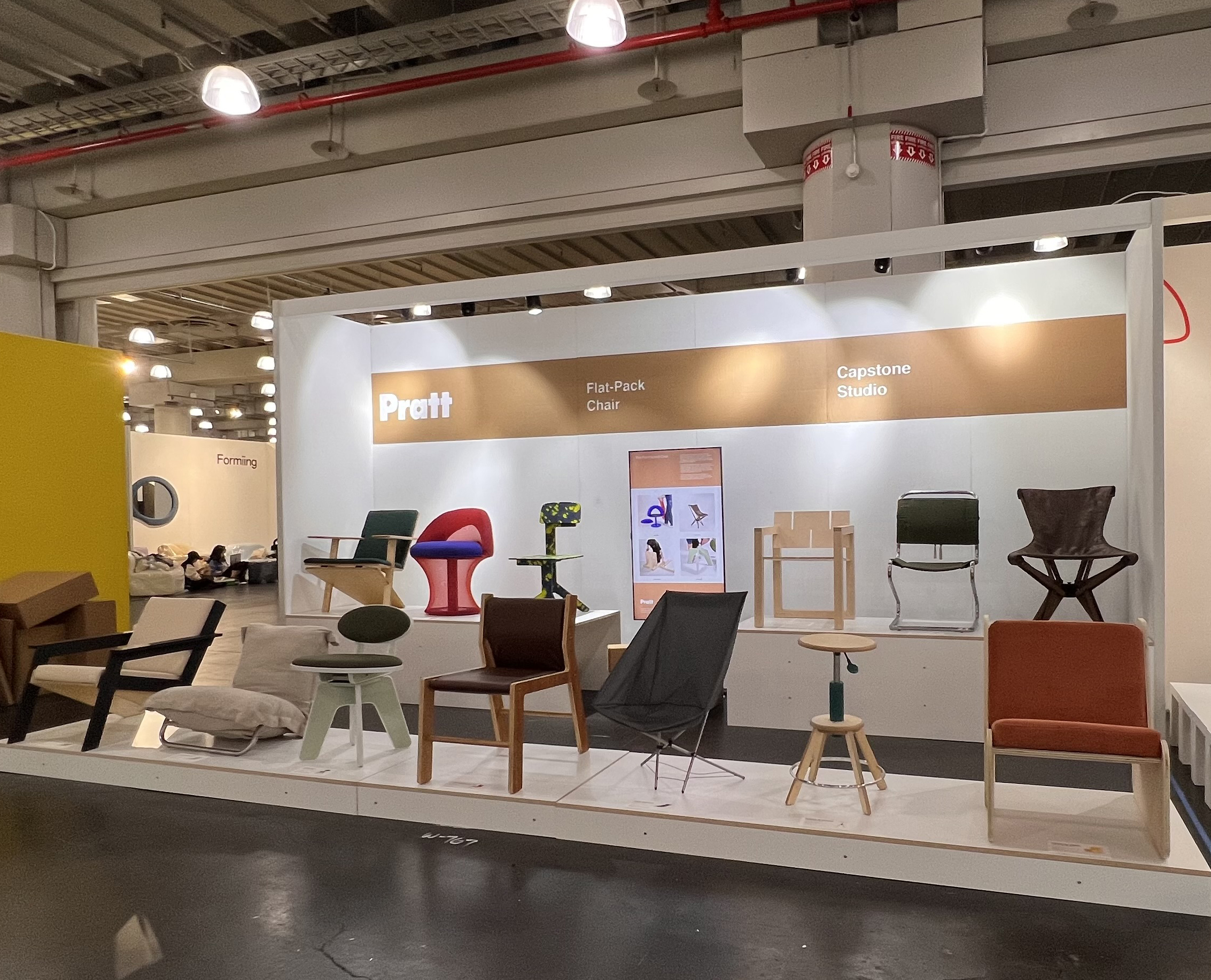 A gallery-style exhibition booth showcases a collection of student-designed chairs under the theme “Flat-Pack Chair” by the Pratt Capstone Studio. Eleven uniquely styled chairs are arranged on a raised white platform, each demonstrating innovative materials, colors, and construction techniques. The backdrop features a brown and white Pratt-branded panel, along with an informational poster. The space is well-lit with industrial overhead lighting, and other exhibits are visible in the background.