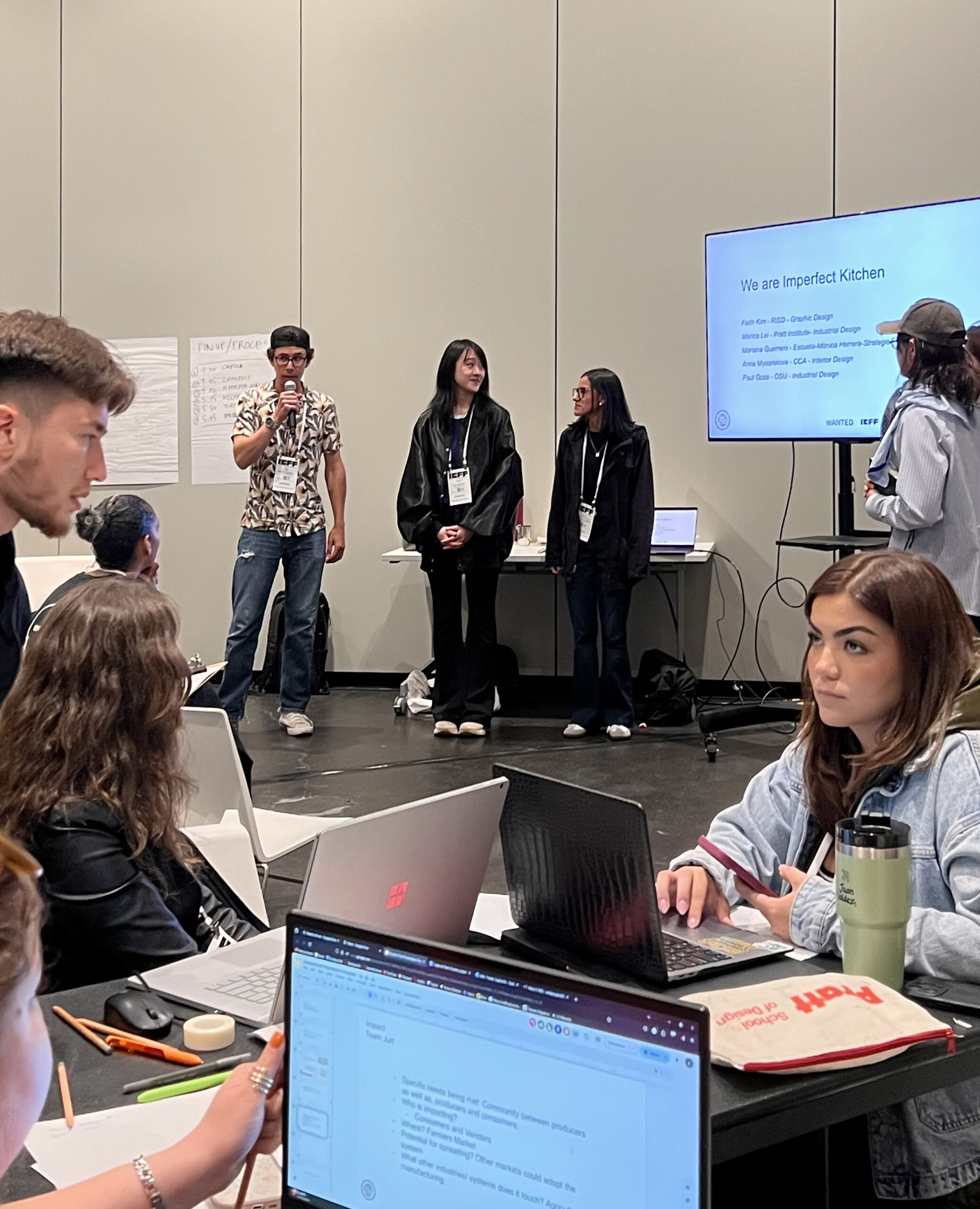A group of students presents a project titled “We are Imperfect Kitchen” in a modern classroom or conference setting. One student speaks into a microphone while three others stand beside him, facing the audience. A screen behind them lists team members' names and design disciplines. In the foreground, other students sit at tables with laptops open, taking notes and working. A visible laptop screen displays a slide with bullet points, and a canvas pouch labeled “Pratt” lies nearby. The atmosphere is collaborative and focused.