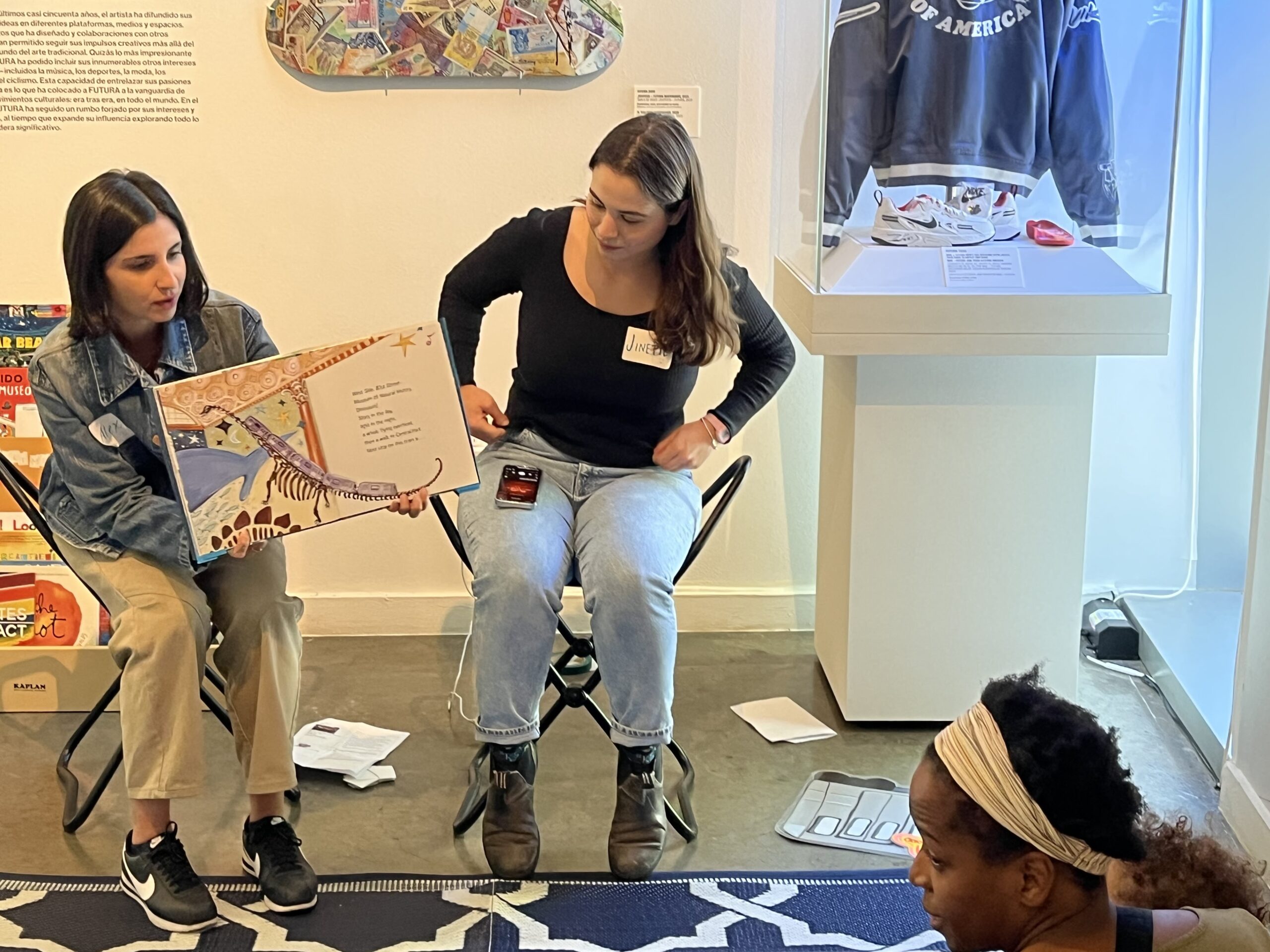 Two women sit in chairs and read a children's book to an audience sitting on the floor.