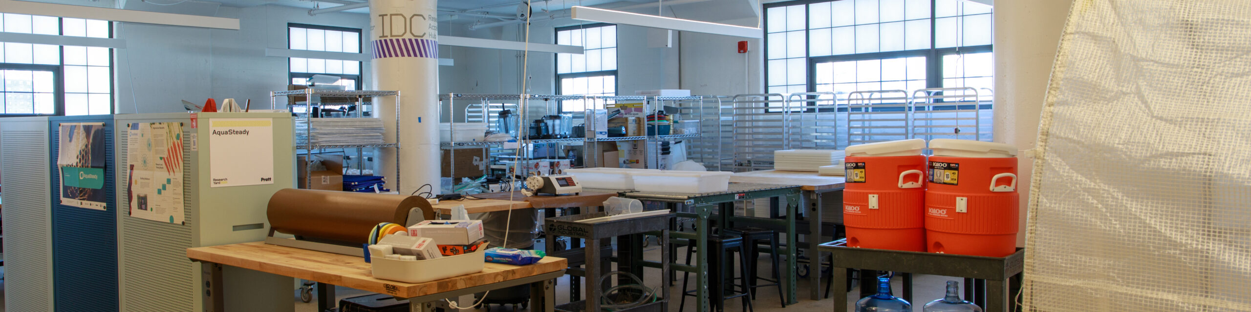 A wide-angle view of a well-lit industrial design or fabrication lab with multiple workstations, high stools, and shelving stocked with tools and materials. A large column labeled “IDC Research Access Hub” stands at the center, and posters for “AquaSteady” are visible on display boards. Two orange Igloo coolers sit on a side table to the right, and grid windows provide natural light across the space.