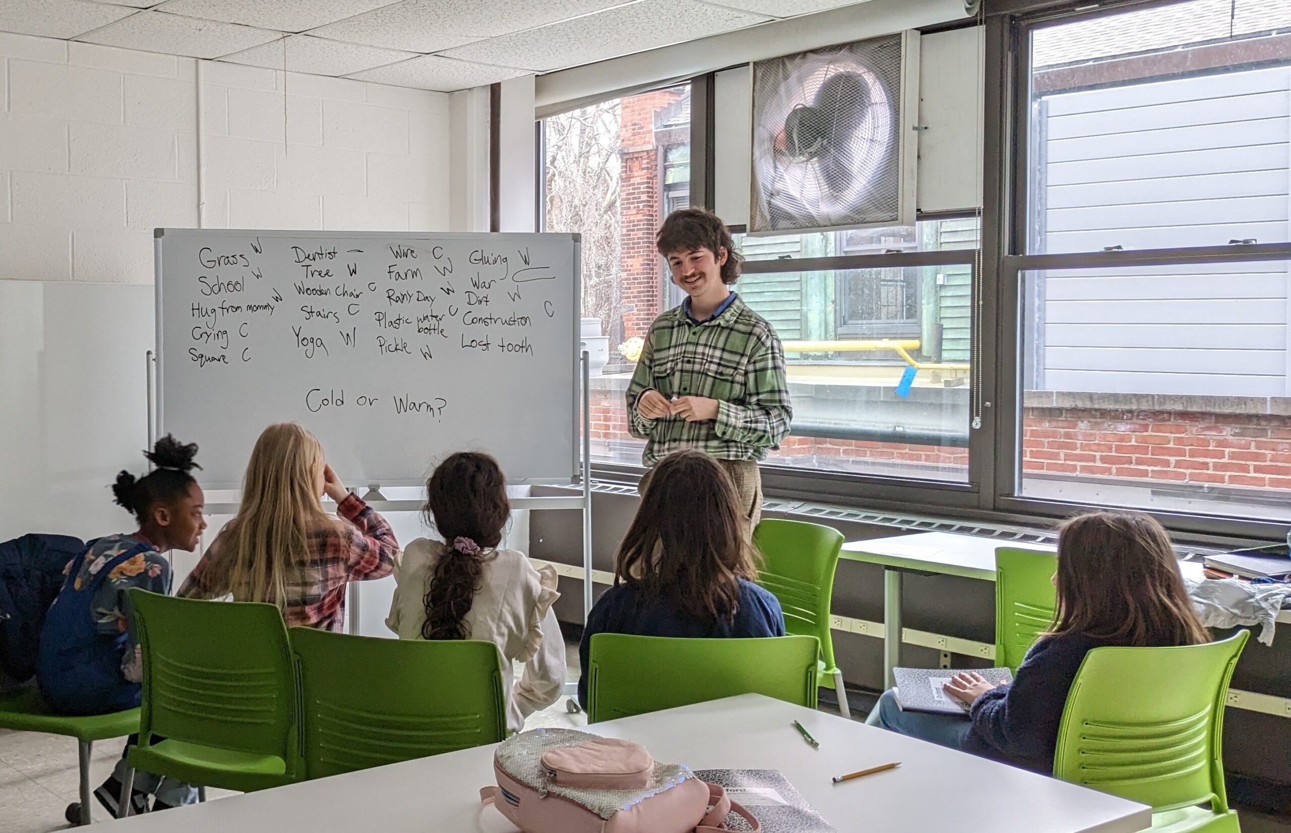 A teacher stands beside a whiteboard with a list of objects labeled “Cold” or “Warm.” Six elementary-aged students sit in green chairs facing the board, engaged in a lesson inside a classroom with large windows showing brick buildings outside.