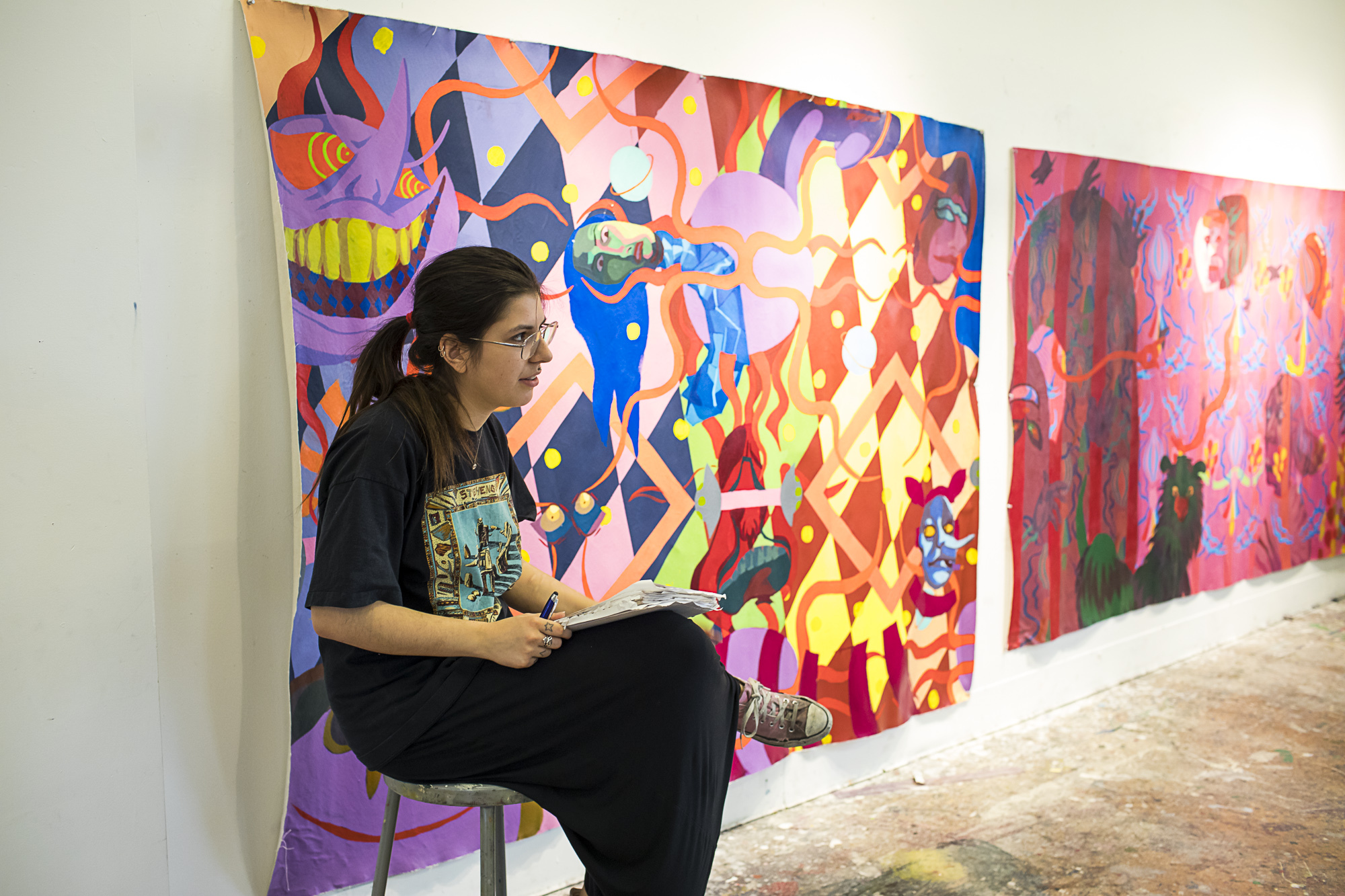 A student sites on a stool in front of two large paintings hung on the wall.