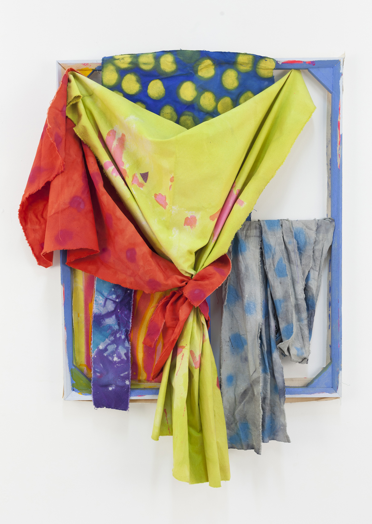 A mixed-media artwork composed of brightly dyed fabrics in red, lime green, purple, and grey, draped and tied around a wooden frame. The background fabric features a bold pattern of yellow circles on a blue field, while the lower layers show paint splatters and textures. The work merges textile and painting traditions, evoking both domestic and sculptural associations.