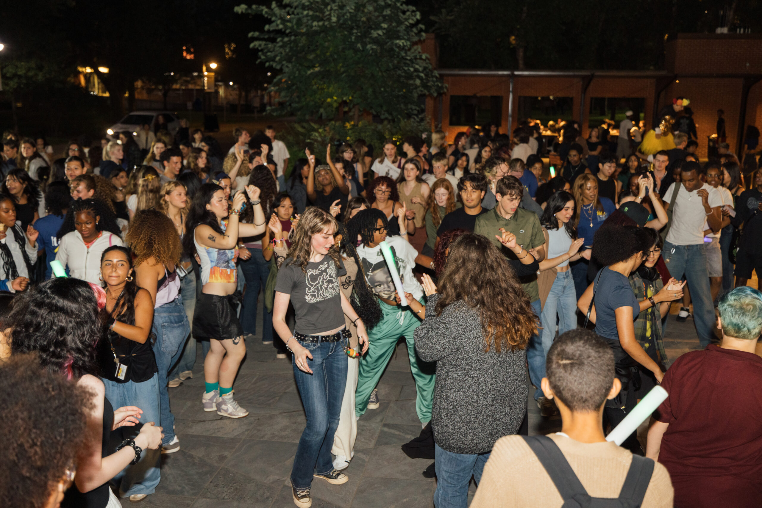 College students dance and mingle in a stone courtyard during the nighttime as the light from a camera flash illuminates the scene.