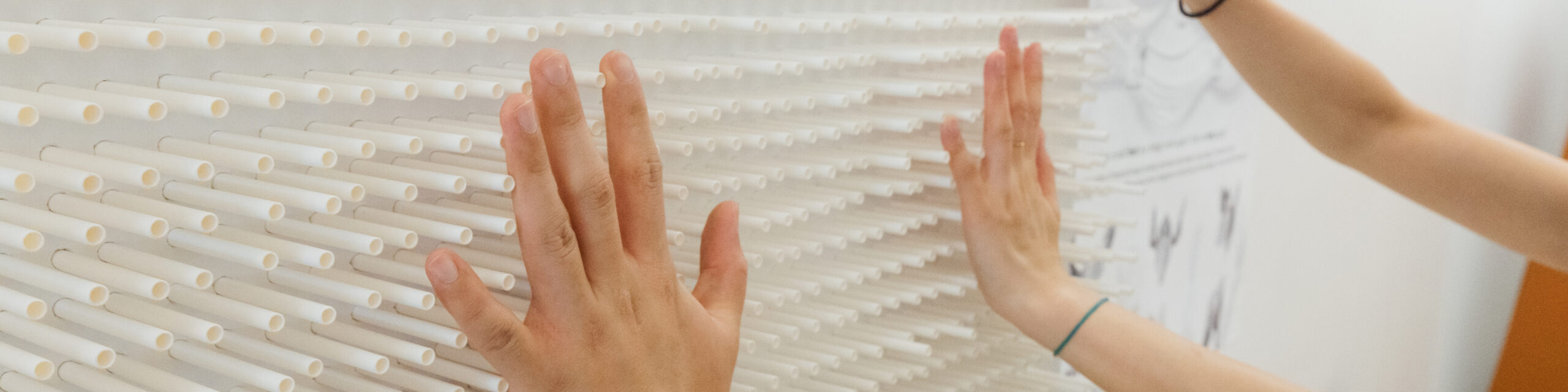Two people engage with a tactile wall composed of evenly spaced white cylindrical rods, which protrude horizontally from the surface. Their outstretched hands press against the rods, creating a sense of interaction or exploration. The wall’s grid-like texture contrasts with the organic motion of the hands. A diagram or poster is partially visible in the background, suggesting a research or design exhibition setting. The image conveys interactivity, sensory experience, and experimental design.