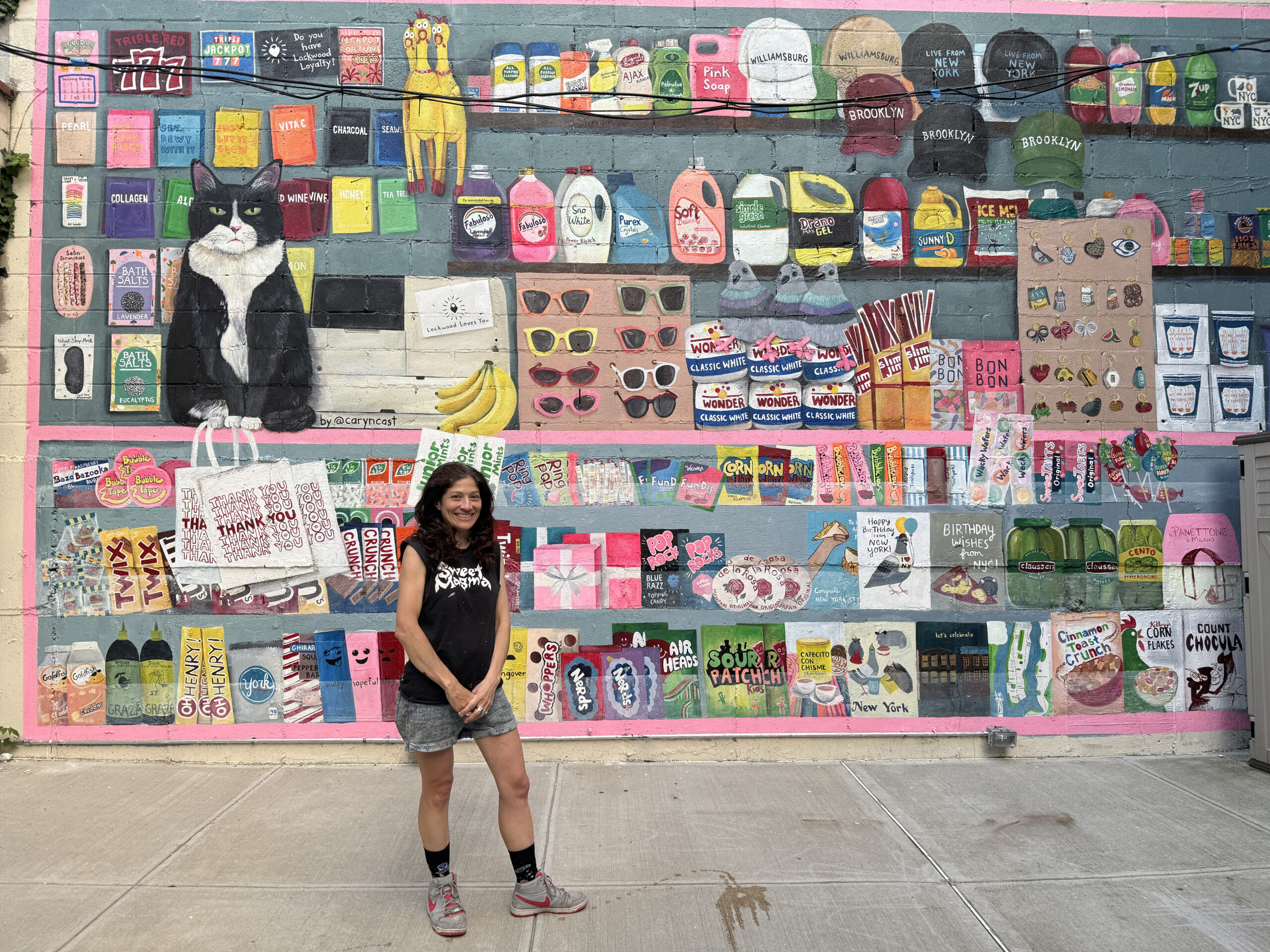 A woman stands in front of a large, colorful mural with images of a cat, shelves, and bodega products.