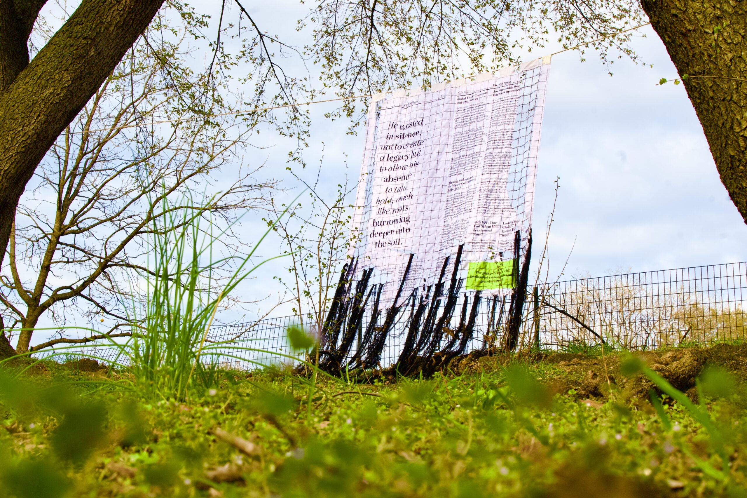 Outdoor art installation featuring a large translucent textile or banner with poetic text, suspended on a mesh frame between trees above a grassy area.