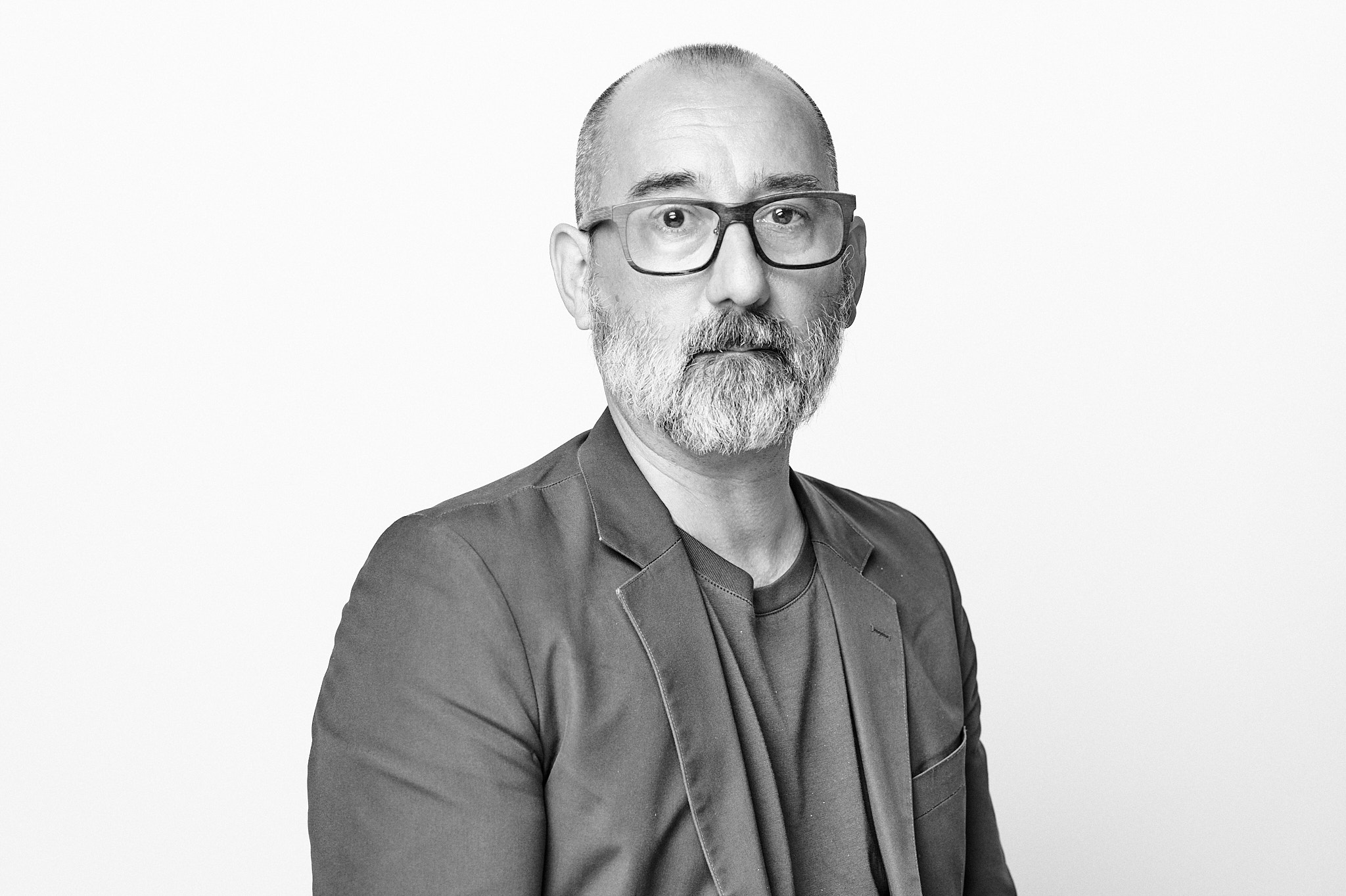 Black-and-white portrait of a man with a close-cropped haircut, beard, and glasses, wearing a blazer over a T-shirt. He sits facing the camera with a neutral expression against a plain white background.