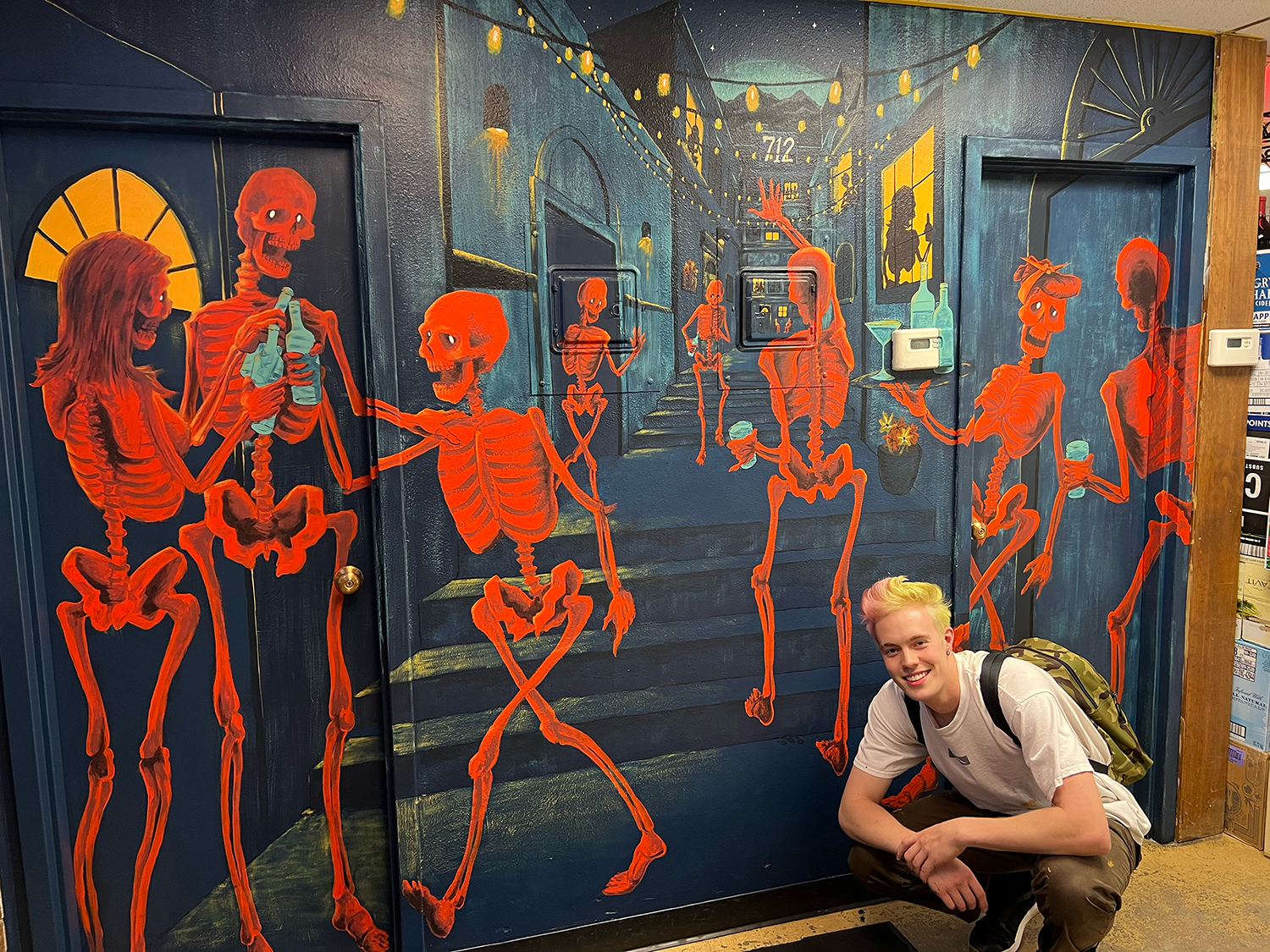 A colorful mural featuring glowing red skeletons socializing and drinking on a nighttime city stoop, with a smiling person crouching in front of the artwork.