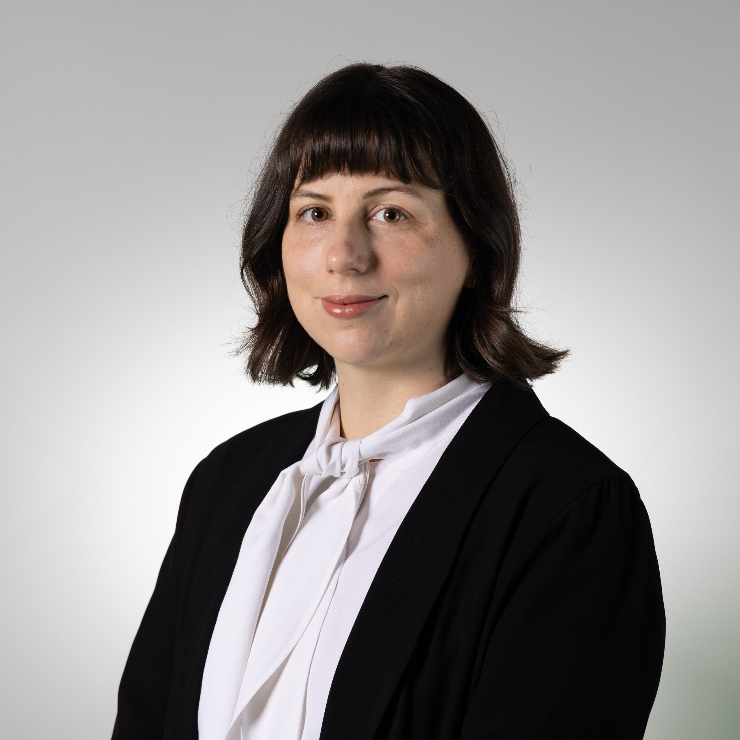 A woman with shoulder-length dark hair and bangs is wearing a black blazer over a white blouse with a bow tie collar. She has a neutral expression, standing against a light gradient background that transitions from white to gray.