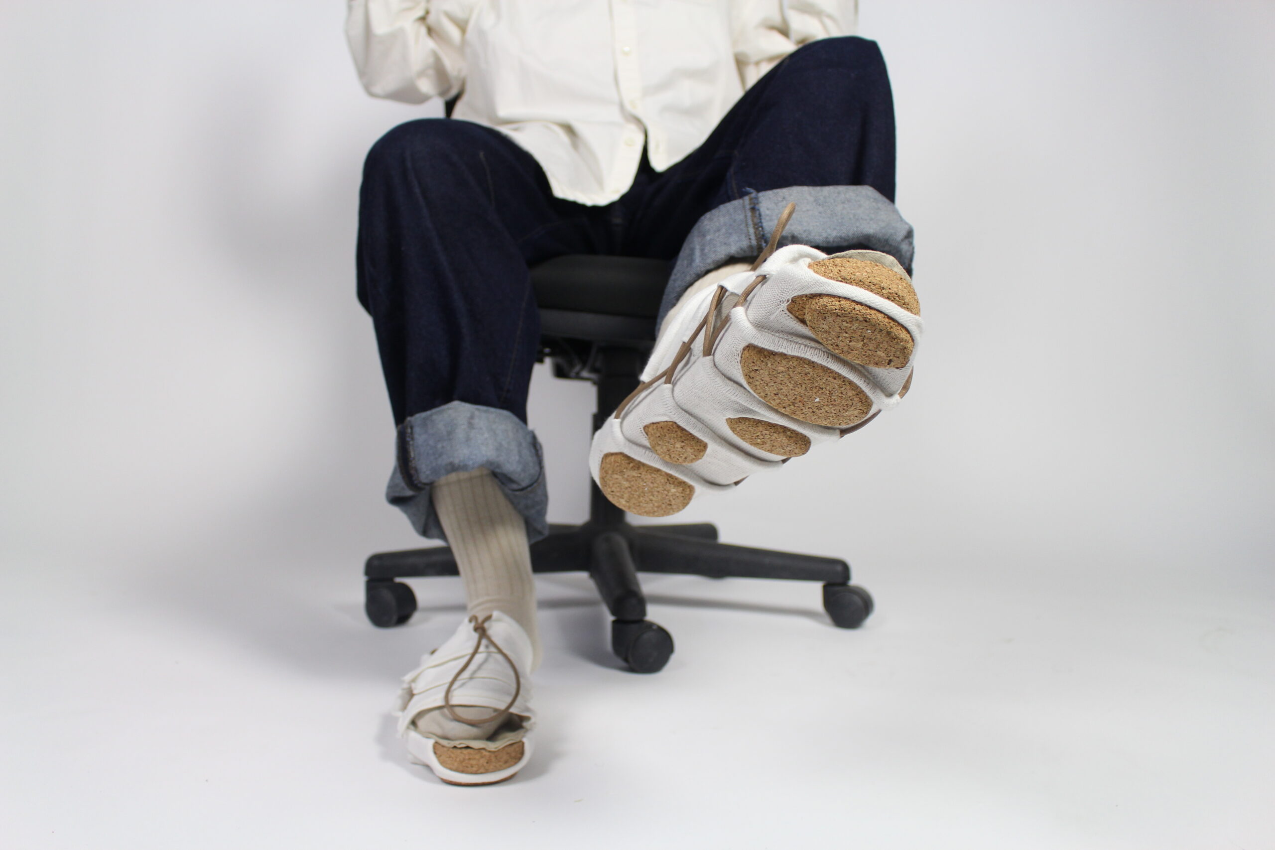 Person seated on a chair wearing uniquely designed sandals with segmented cork soles and white fabric uppers, one foot lifted to show the sole.