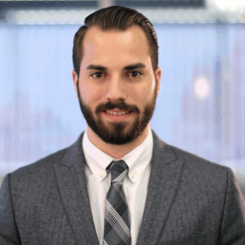 A professional headshot of a man wearing a dark gray suit jacket, white dress shirt, and gray plaid tie. He has neatly styled dark hair parted to the side and a well-groomed beard. He is standing against a softly blurred background featuring vertical blinds and natural daylight, giving the photo a formal, corporate tone. The subject faces the camera directly with a neutral, confident expression and a faint smile.