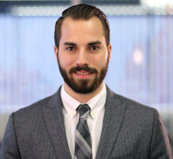 A professional headshot of a man wearing a dark gray suit jacket, white dress shirt, and gray plaid tie. He has neatly styled dark hair parted to the side and a well-groomed beard. He is standing against a softly blurred background featuring vertical blinds and natural daylight, giving the photo a formal, corporate tone. The subject faces the camera directly with a neutral, confident expression and a faint smile.