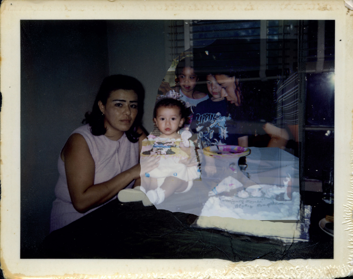 A faded, polaroid-style photograph showing a celebration. In the foreground, a woman with dark hair and a sleeveless pink top is sitting beside a baby who is dressed in white and sitting on a table. Behind them, two children and another adult are gathered around a cake that has colorful decorations. The background includes windows with blinds partially open, letting in light. The overall image has a nostalgic feel with some scratches and wear visible.