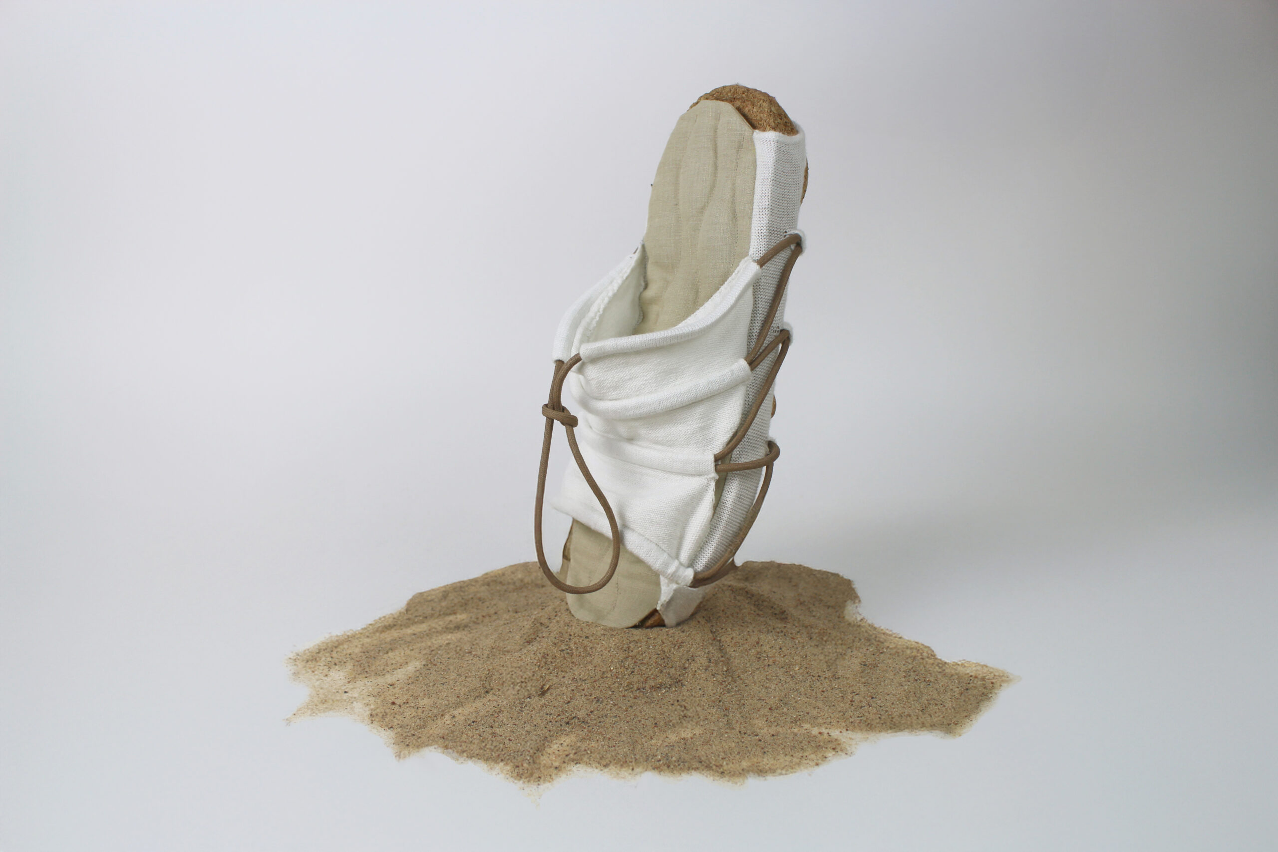 Single sandal standing upright in a mound of sand, featuring a cork sole, white fabric upper, and brown leather lacing.