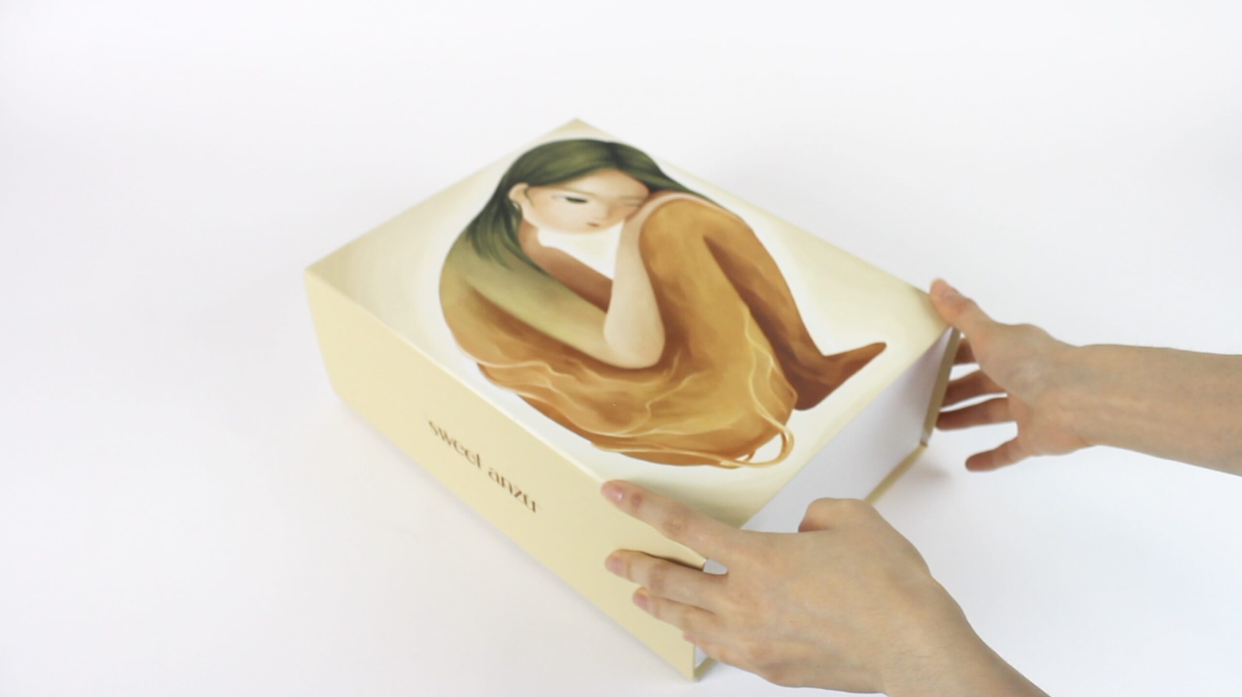 A person is shown holding a yellow box with an artistic illustration on the top. The illustration features a figure sitting with their knees hugged to their chest, displaying long hair and a contemplative expression. The box has the text